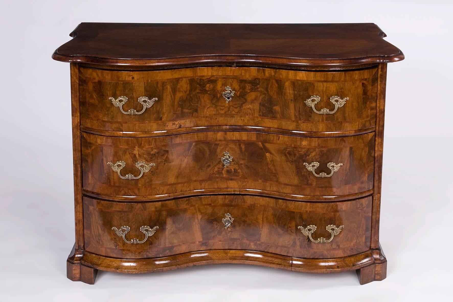A walnut wood Baroque commode from the Hessian state of Germany. A three-parted dresser with a wonderfully curved front. It is made of walnut wood and walnut-grain with a mirrored veneer on the cover plate. The locks are originals but the fittings,
