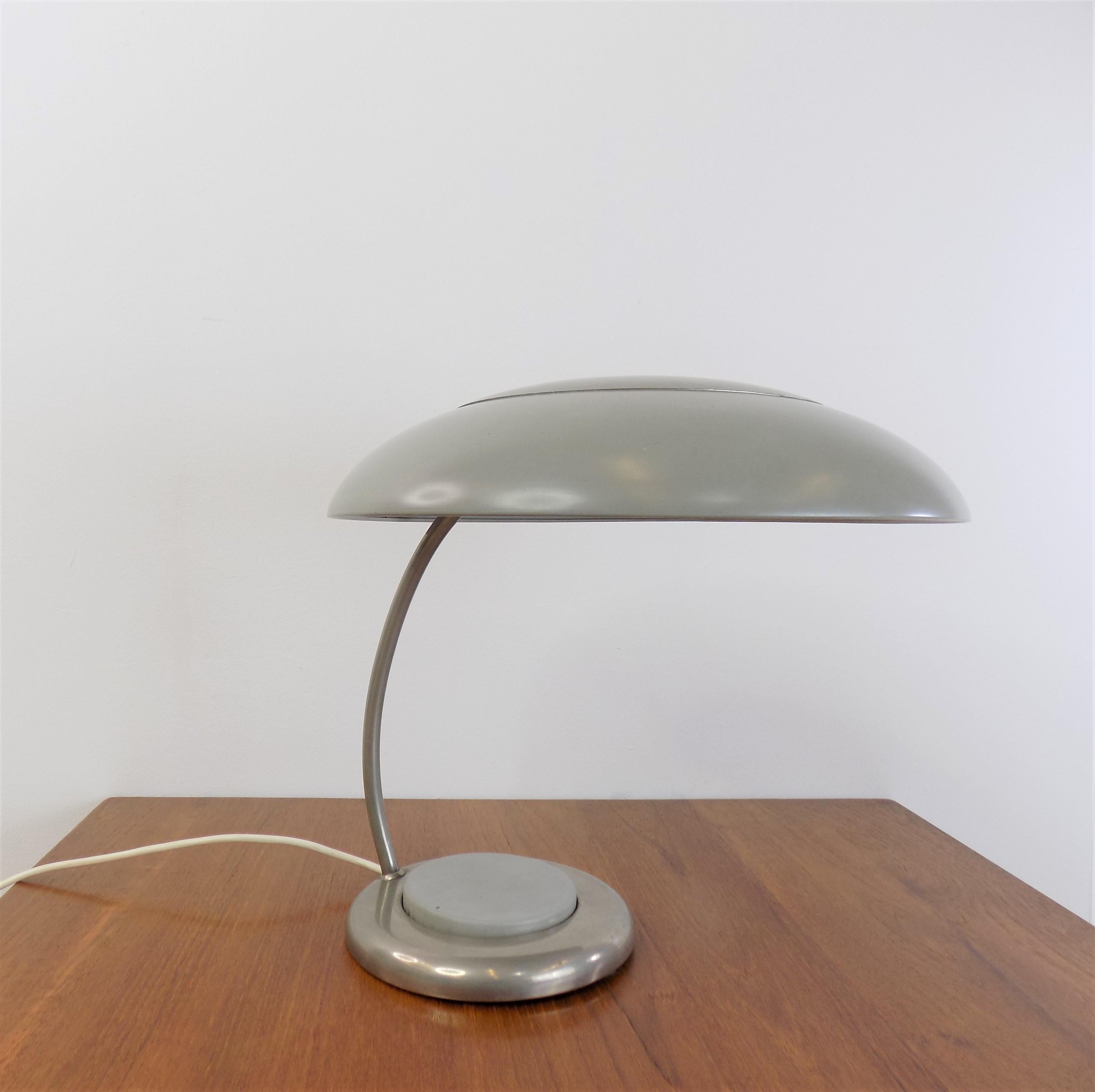 La lámpara de mesa está en muy buen estado, con una atractiva pátina. La pantalla dominante de metal grisáceo y cubierta cromada presenta mínimos signos de desgaste,  así como la base de la lámpara, que impresiona por el interruptor redondo