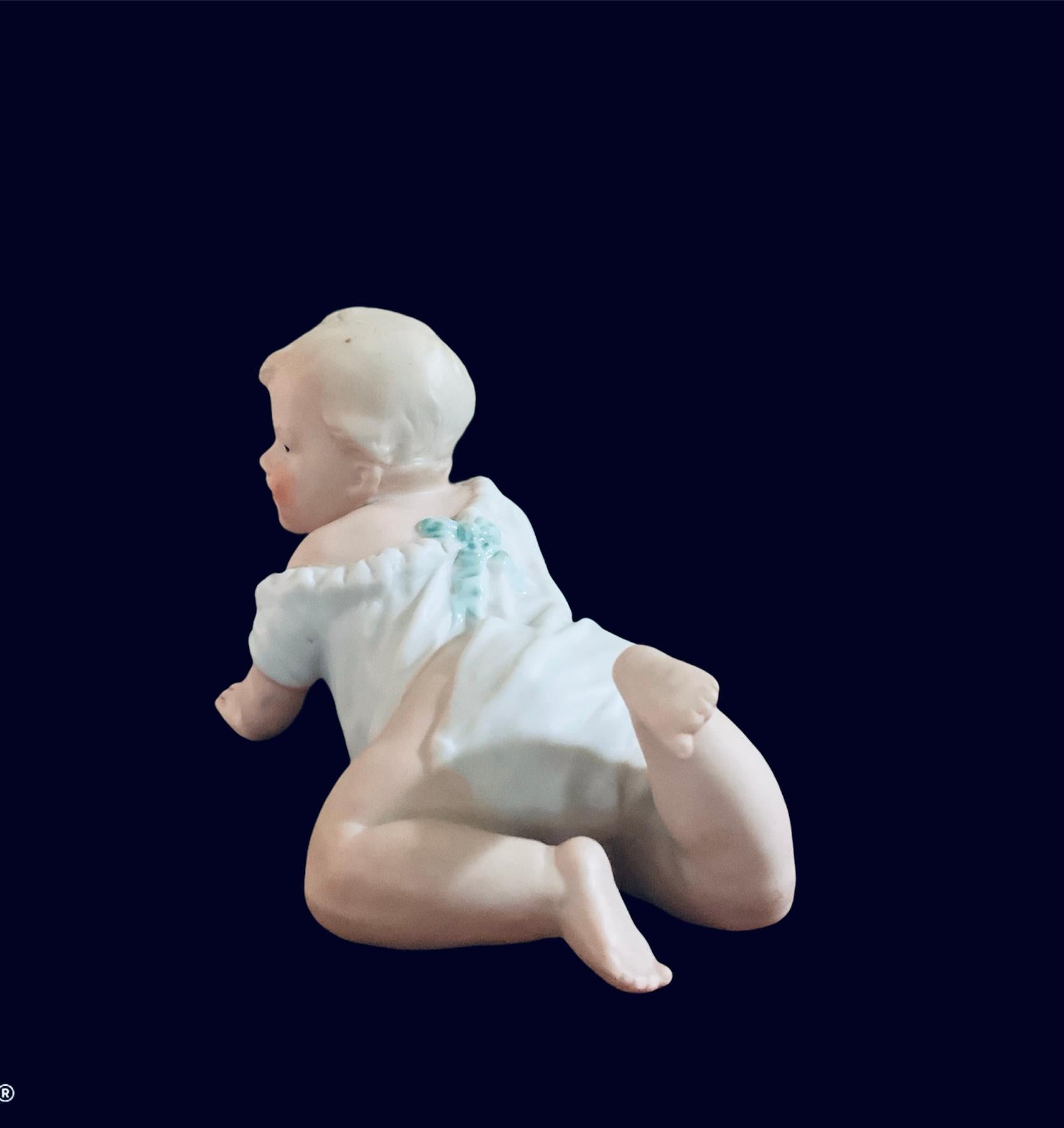This is a bisque porcelain piano baby boy figurine. He is crawling and looking kind of surprised to the side with a shy smile. He is wearing a delicate long white chemise with ruffles, large blue bow and ribbons in the back. His hair is curly