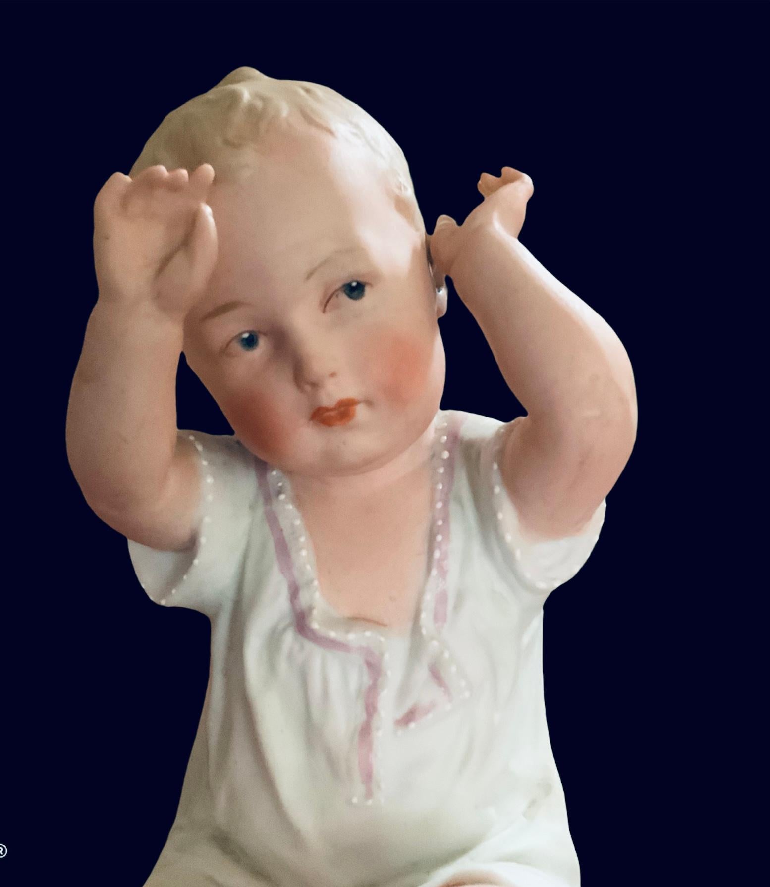 This is a bisque porcelain piano baby girl figurine. She is seated crossing her legs with her arms up and wearing a delicate white chemise with pearly buttons and pink ribbons. Her hair is curly blonde, her cheeks are pinky and her eyes are blue. A