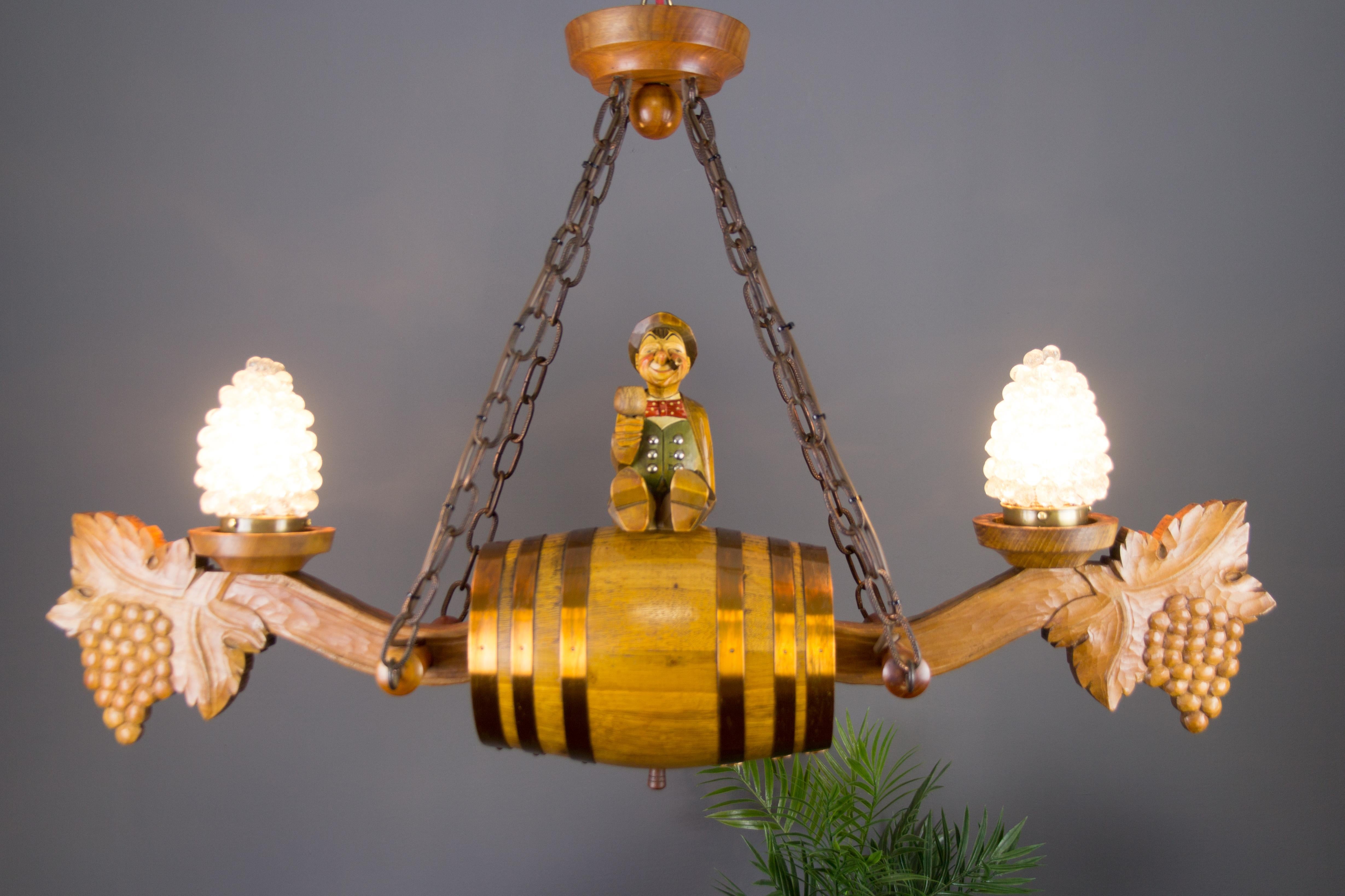This interesting and unusual hand-carved wooden chandelier features a centered large wine barrel and a wooden figurine of the winegrower sitting on it. The ends of the chandelier are decorated with two impressive carved bunches of grapes and two