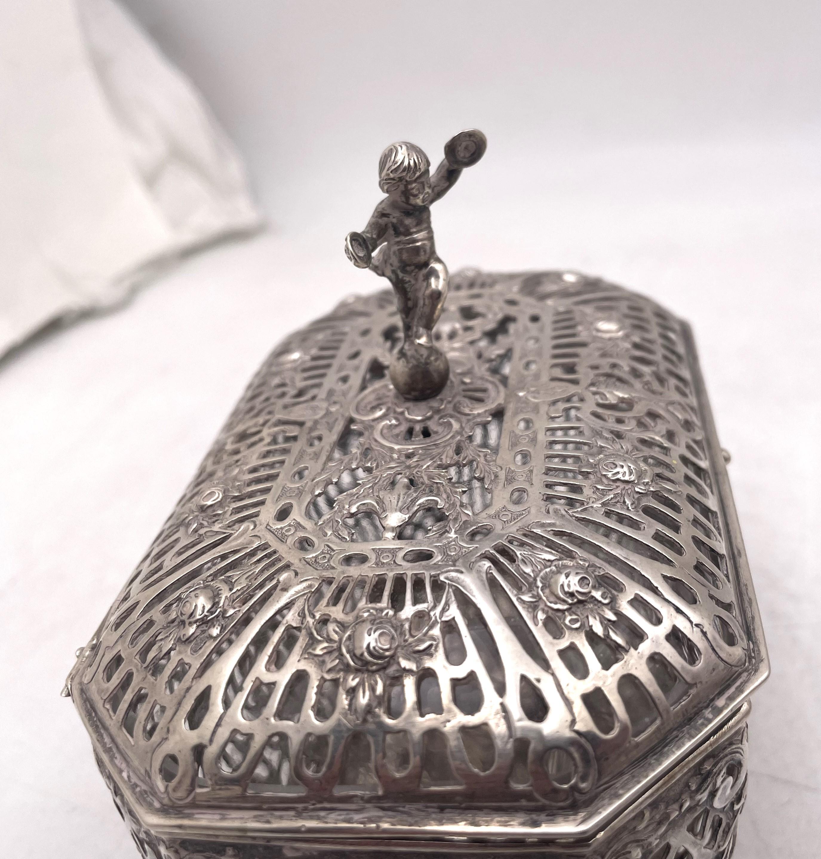German, continental 0.800 silver casket, beautifully adorned with a putti on top, with pierced geometric and floral motifs. There is glass inside the top part of the casket. It measures 5 1/3'' in length by 3 1/2'' in width by 4 3/4'' in height and