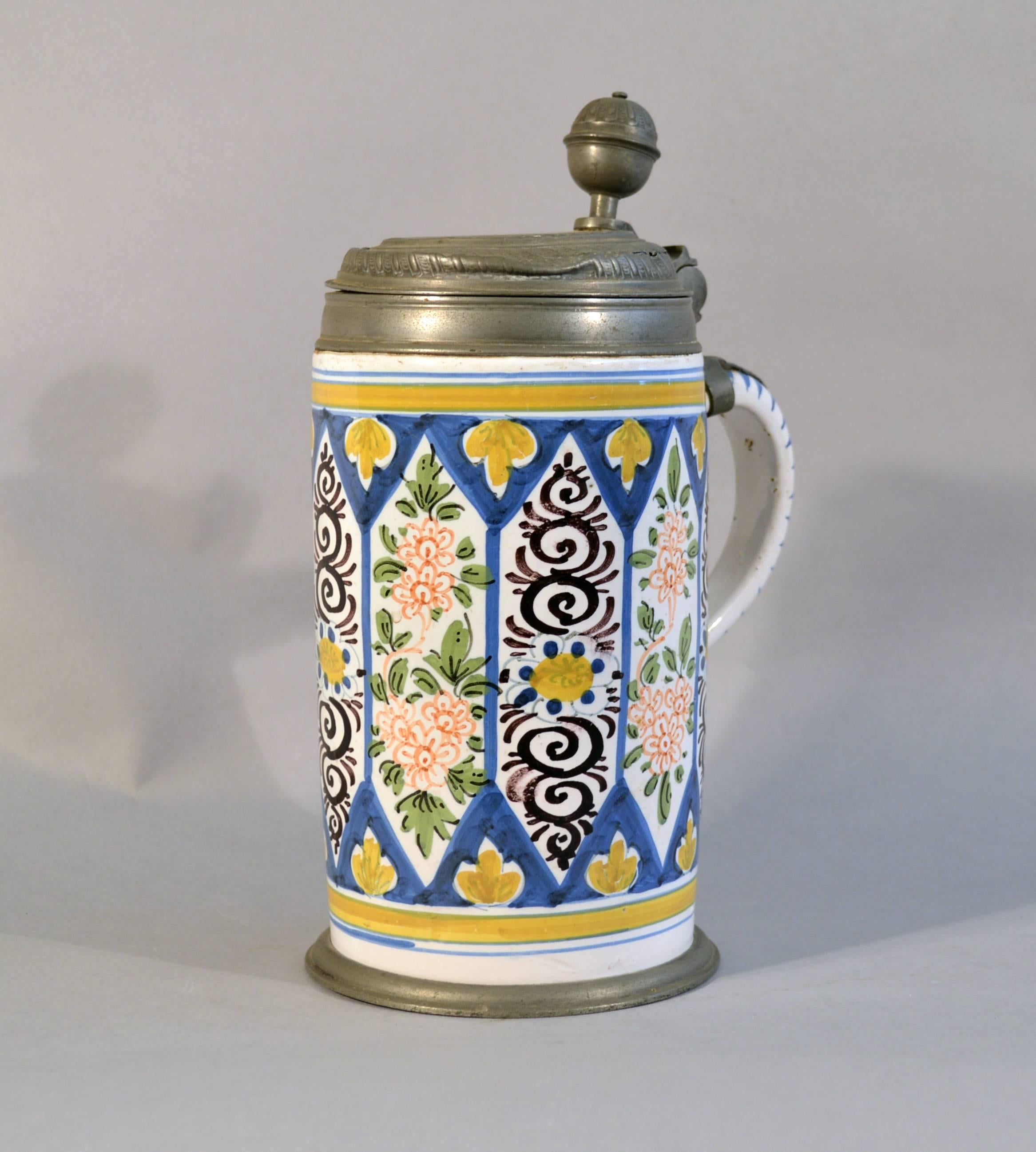 German Faience (Tin-glazed earthenware) Pewter-mounted Tankard with Bands of Flowers, 
circa 1760.

A cylindrical German tin-glazed earthenware tankard with pewter foot and cover. The tankard is painted with vertical panels of alternating