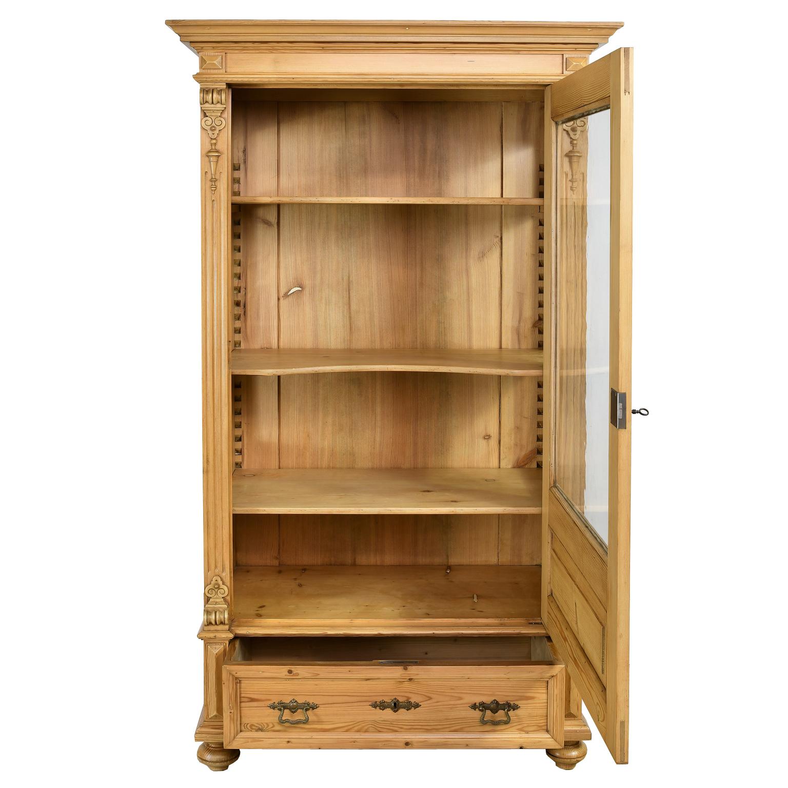 A charming and beautifully-crafted vitrine or cupboard in pine with one door above a storage drawer, and offering a glass panel on the front and sides. Fluted sides and corners are decorated with applied carvings, with moldings around all of the
