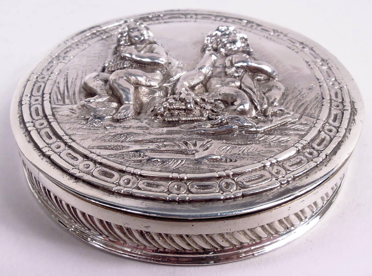 Caja de baratijas rococó alemana de plata de ley, hacia 1930. Redonda con tapa abatible. En la parte superior de la portada, en altorrelieve, hay dos querubines sentados en la hierba: Uno toca el arpa y el otro escucha mientras come uvas. Cenefas