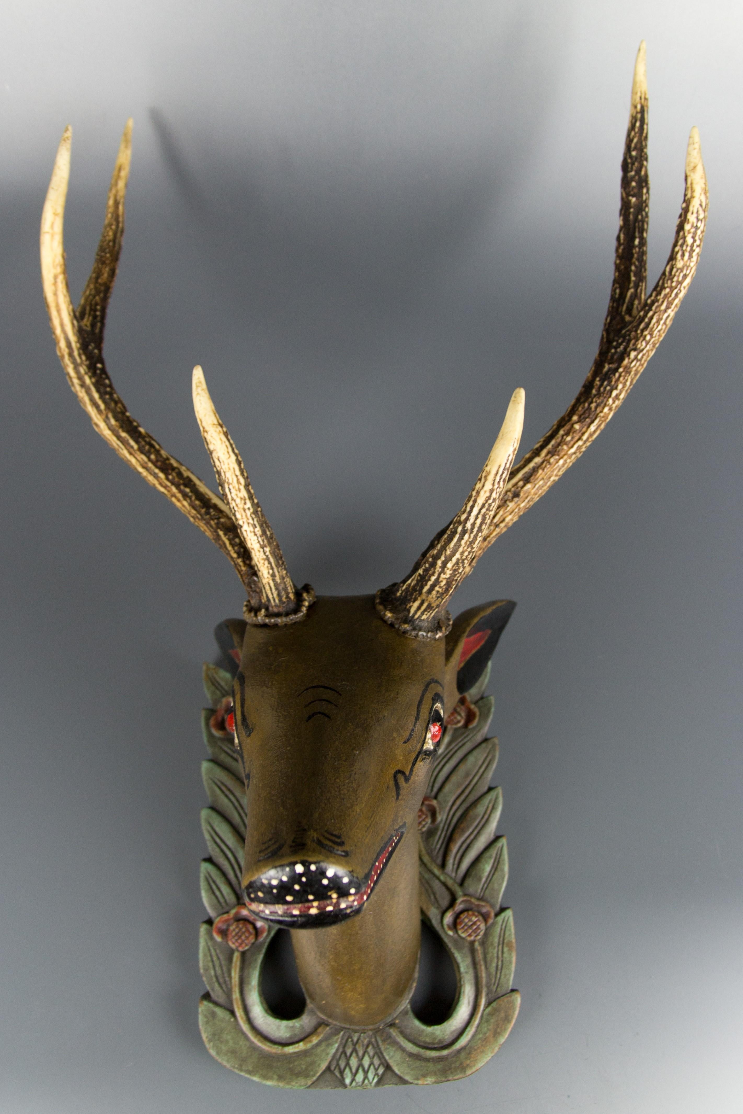 Very unusual and adorable hand carved and hand painted wall hanging deer head with genuine antlers mounted on a carved wooden plaque. The wooden plaque depicts green leaves and red flowers in the Art Nouveau style. The antlers are attached with