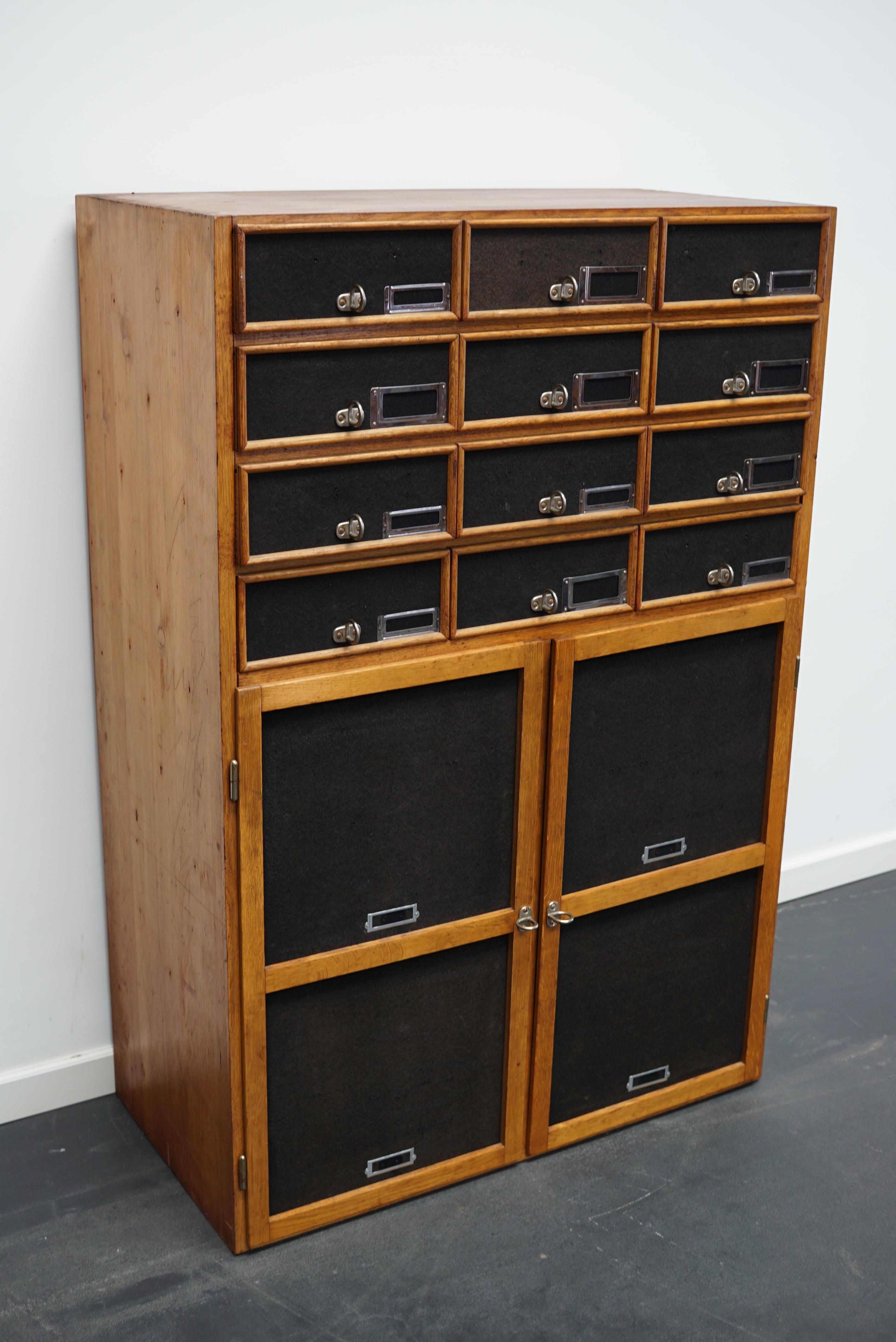 This apothecary cabinet was made circa 1940s in Germany and was later used in a hardware store in Belgium until recently. It features 12 drawers with metal handles / name card holders and two doors at the bottom with two shelves behind them. The