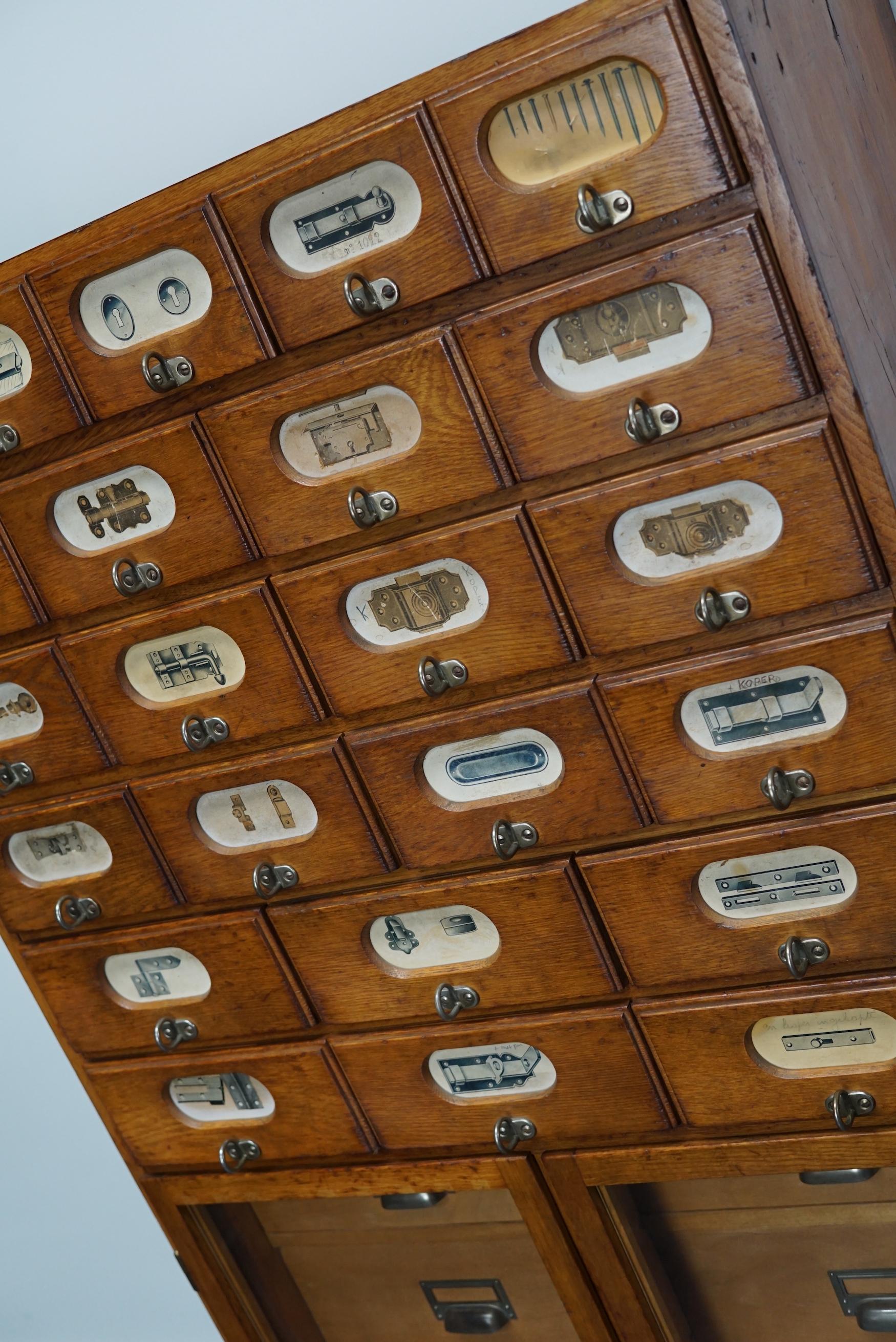 This apothecary cabinet was made circa 1940s in Germany and was later used in a hardware store in Belgium until recently. It features many drawers in different sizes with metal handles and the labels of the contents. It has two doors with glass