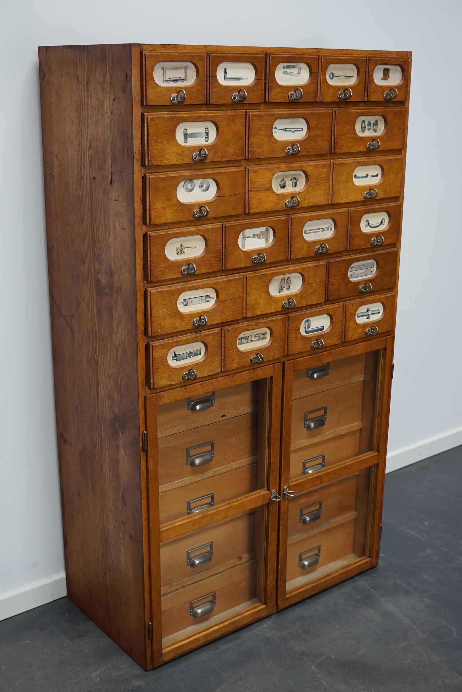 This apothecary cabinet was made circa 1940s in Germany and was later used in a hardware store in Belgium until recently. It features many drawers in different sizes with metal handles and the labels of the contents. It has two doors with glass