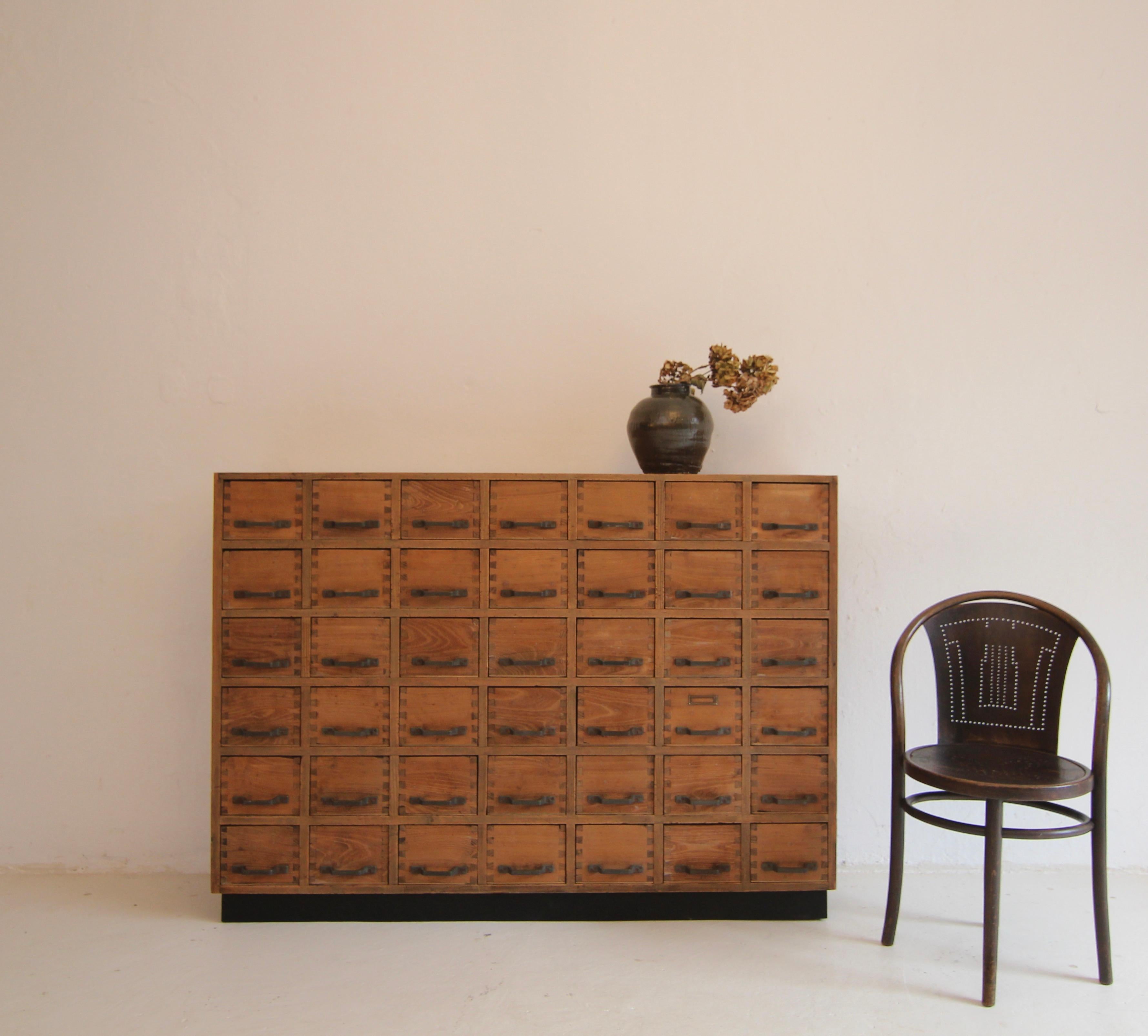 Vintage drawer cabinet from an old cobbler's workshop.

A great unique piece in a purist design with a very beautiful patina.

Pinewood cabinet on a slightly recessed black plinth with a total of 42 dovetailed oak drawers with plywood shelves and
