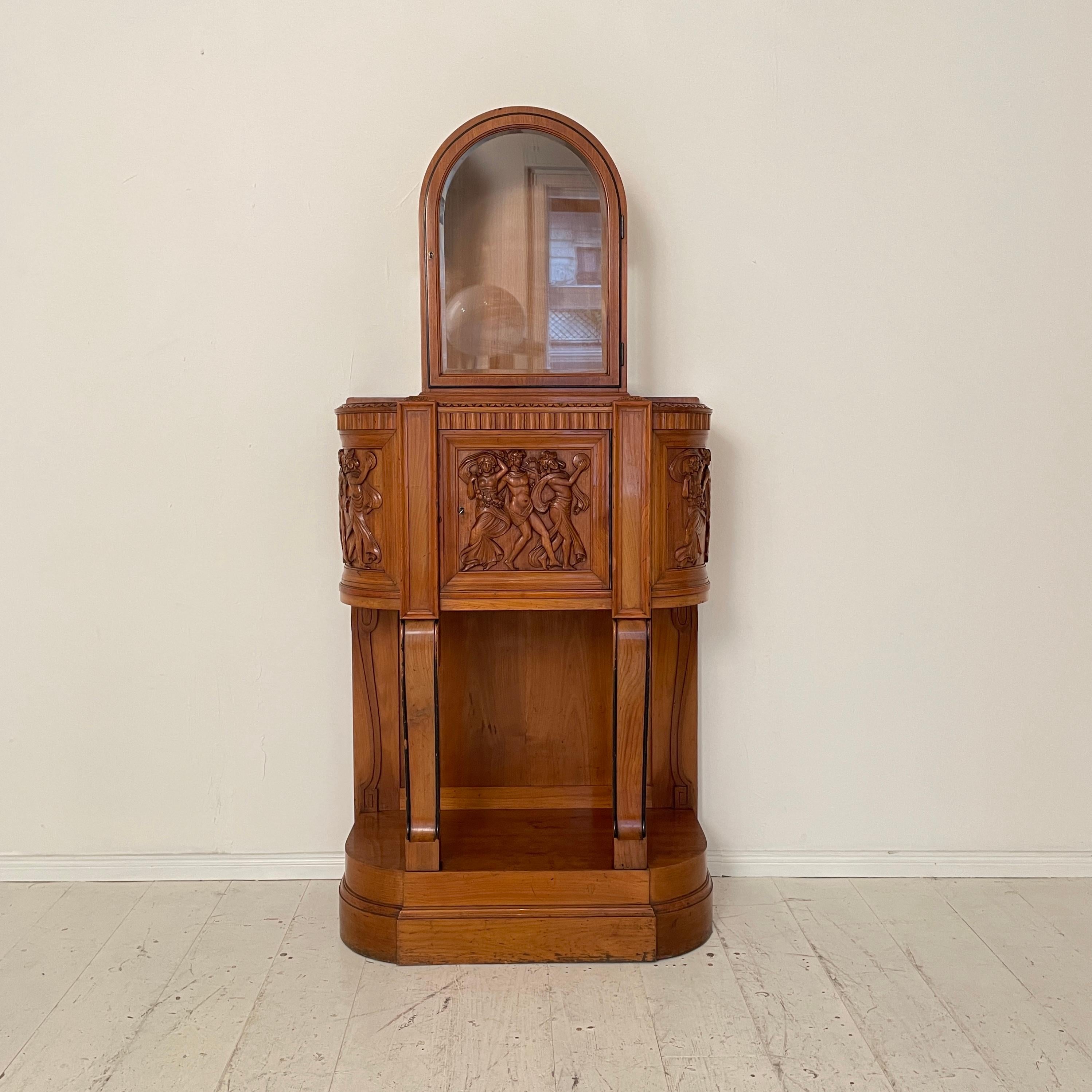 This German Jugendstil cabinet, circa 1905, is a stunning example of the era's artistic flair. Crafted from richly-toned cherry wood, the cabinet's lower half is intricately carved with a lively scene depicting dancing figures celebrating after the
