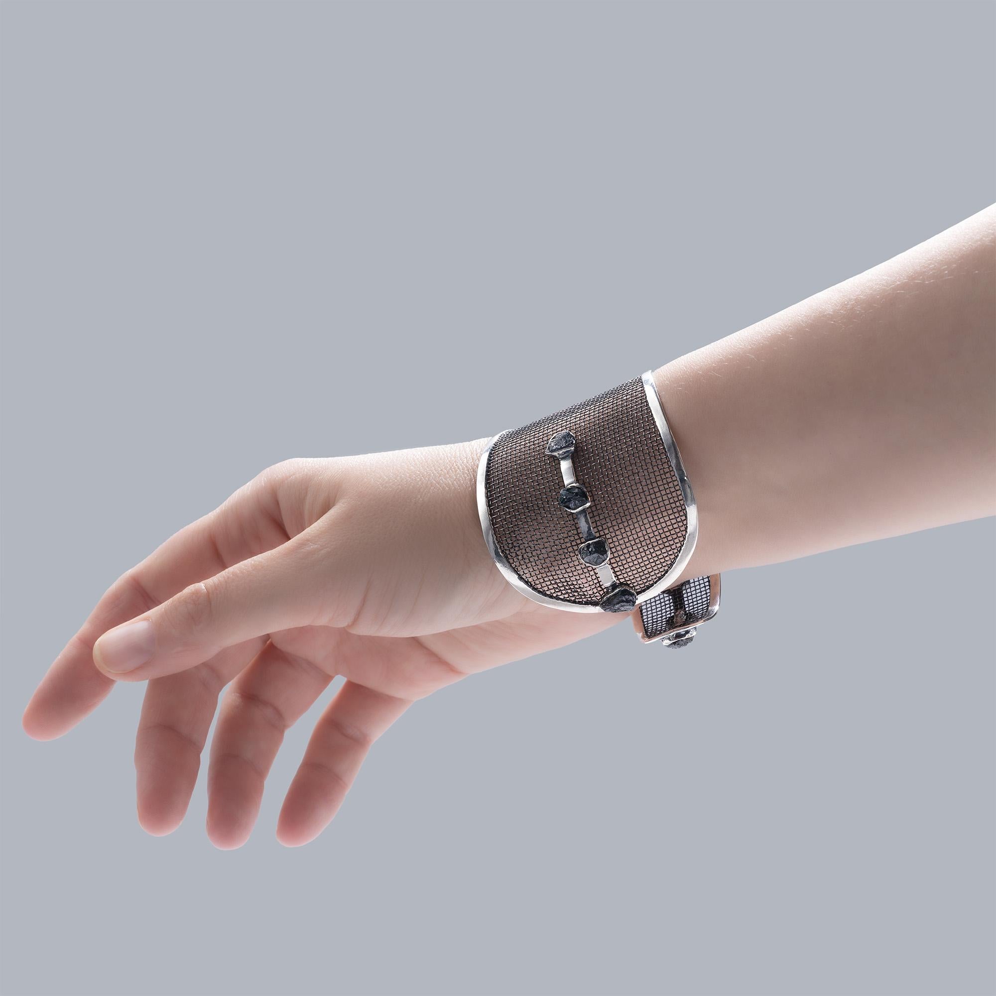 Das Espalier-Armband verbindet industrielle Struktur mit organischer Rohheit. Er ist aus oxidiertem 925er Silber und Titaniumgeflecht gefertigt und fühlt sich sowohl robust als auch edel an. Eine Reihe von rohen, ungeschliffenen Saphirkristallen