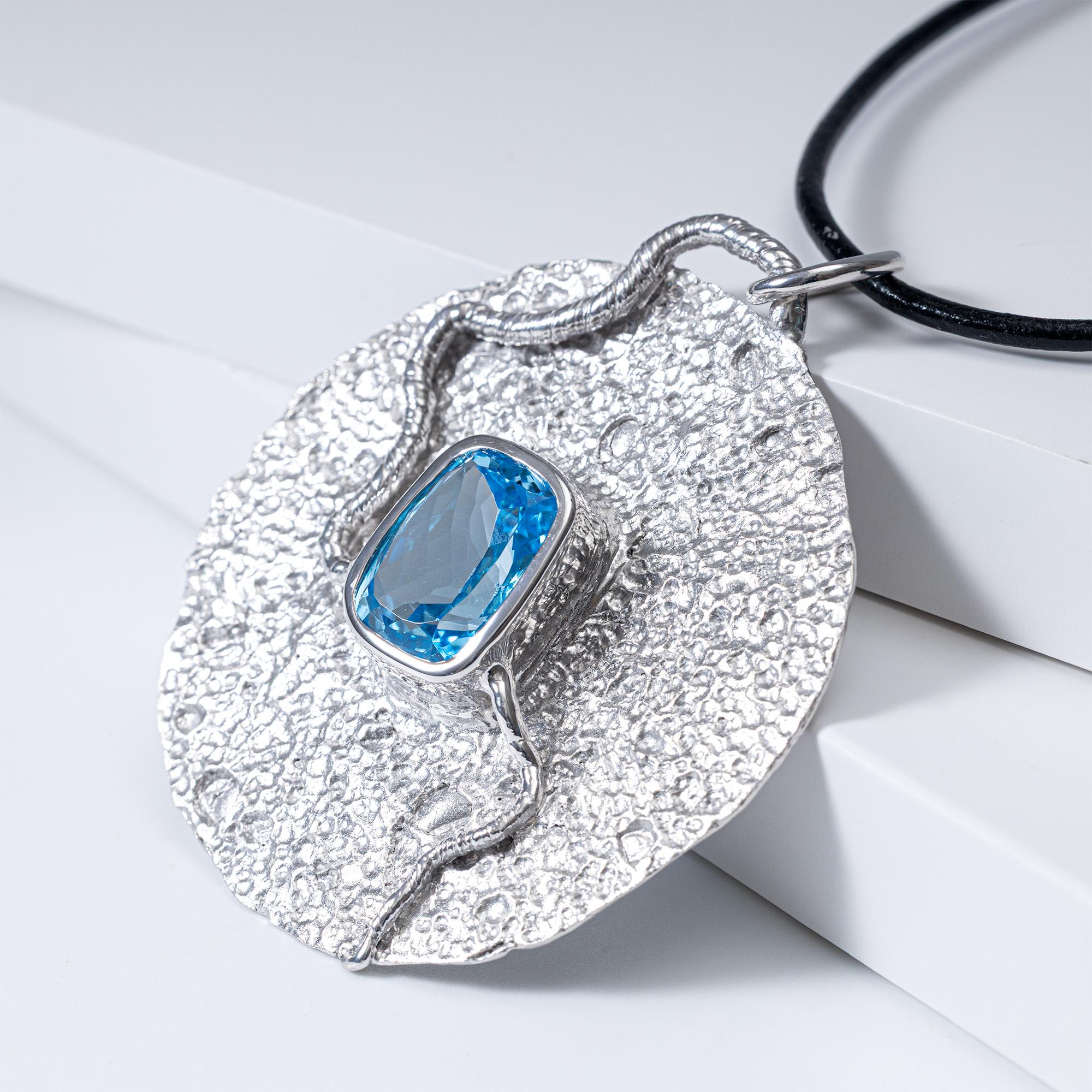 Oread è un ciondolo a forma di disco realizzato a mano in argento 925. Al centro si trova un grande topazio blu tagliato a cuscino, luminoso e trasparente come una sorgente di montagna, incastonato in modo da far passare la luce attraverso la