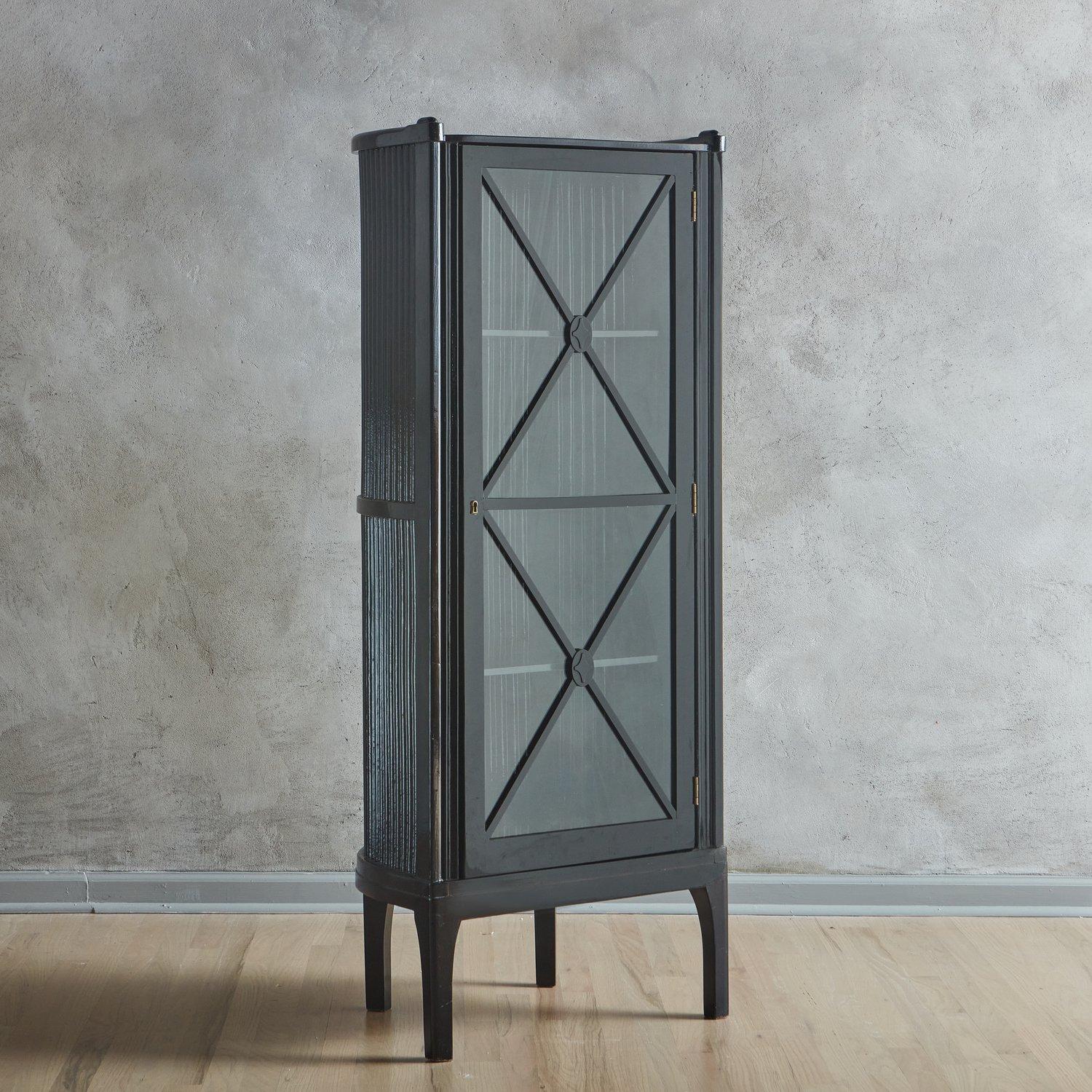 An early 20th Century cabinet designed by Hans Gunther Reinstein featuring a black lacquered wood frame with a fluted barrel back. This elegant piece has a hinged glass door with two shelves and stands on tall block legs. Retains partial ‘Ellmauer