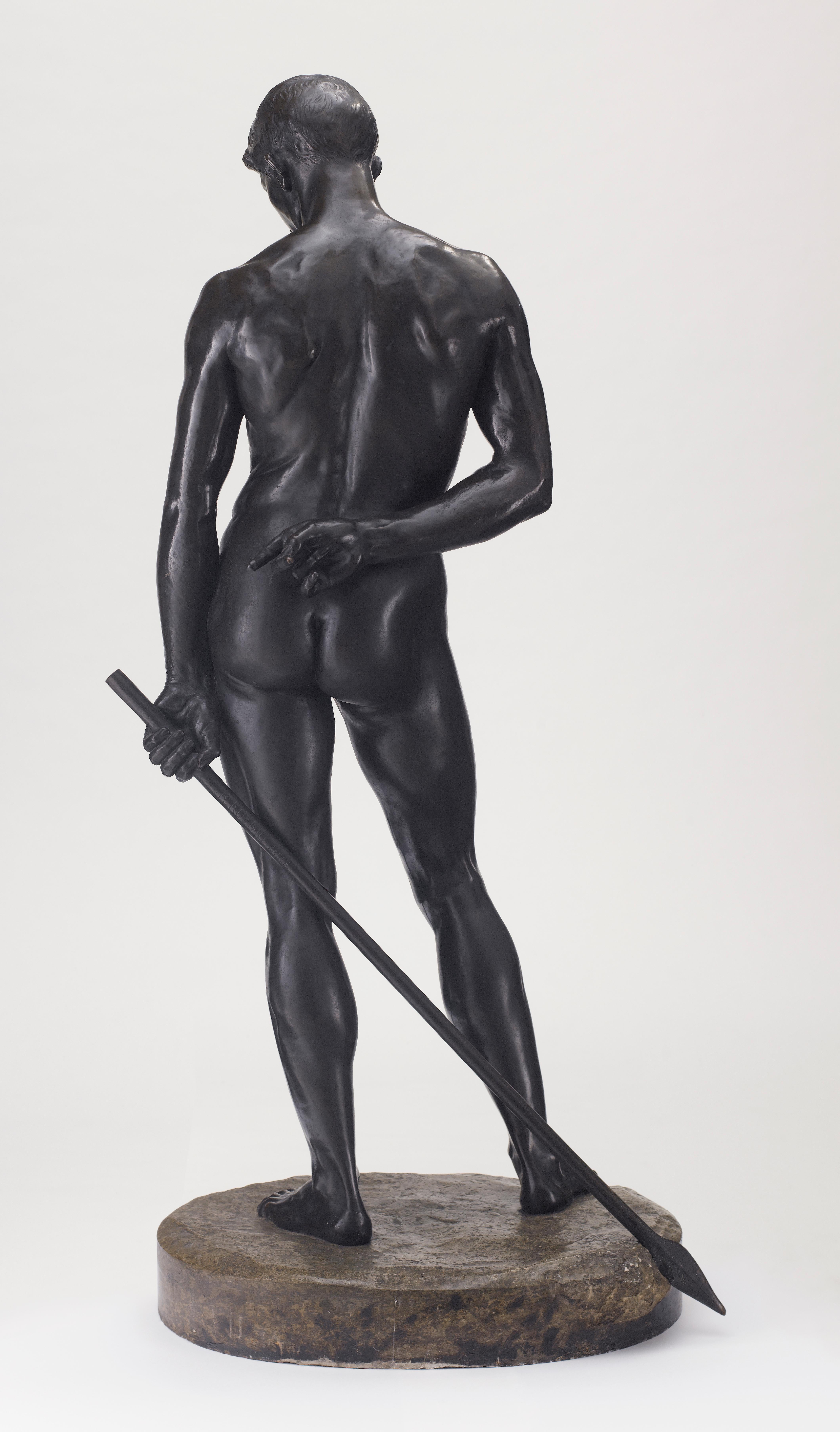 Life-size bronze sculpture, dark patina, mounted on an oval, naturalistically modeled bronze plinth, imitating a stone base. Standing male nude, holding a spear in his left hand, his right clasped behind his back. Signed and inscribed on the lance: