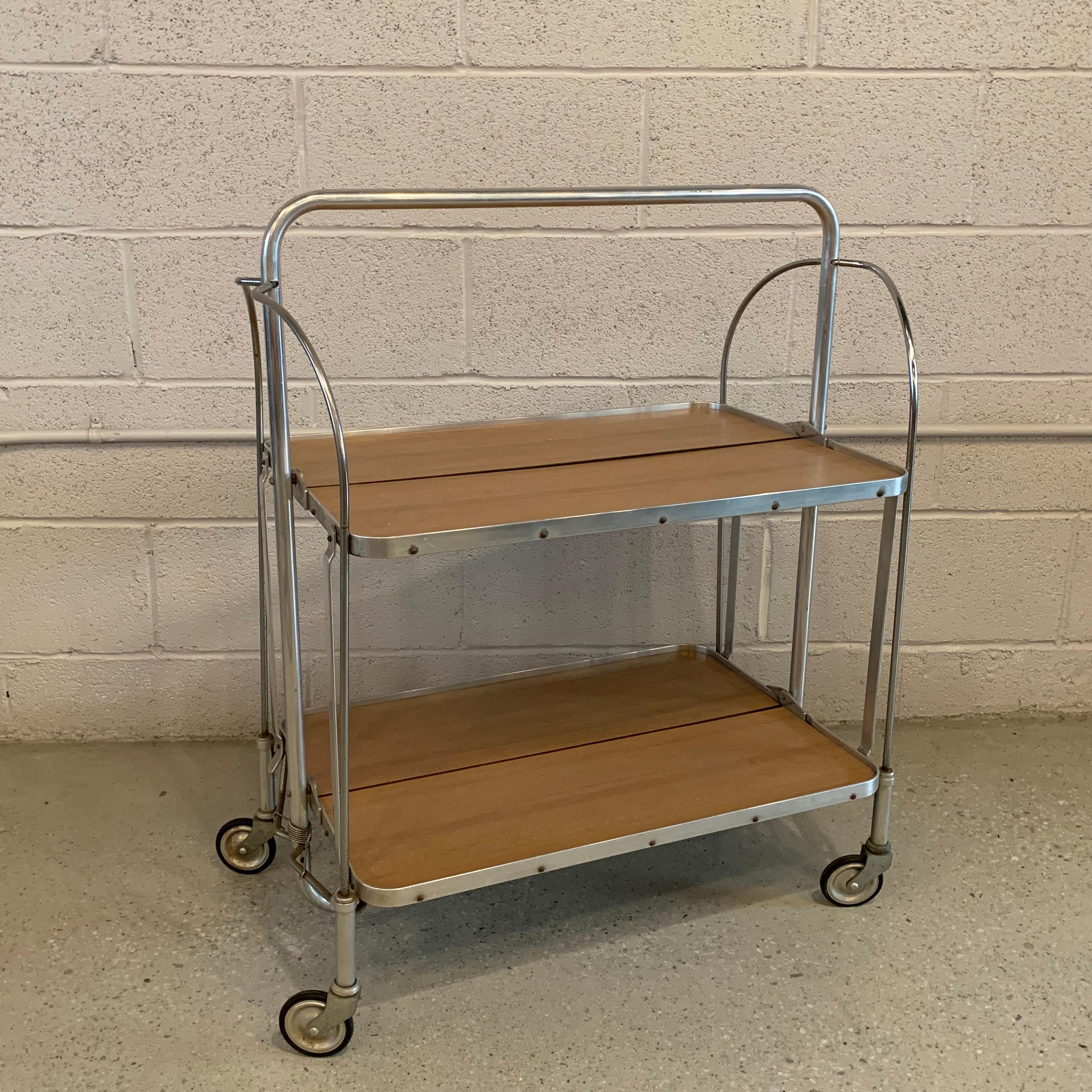 German, Mid-Century Modern, rolling, serving cart by J. Dienstag features a tubular aluminum frame with faux wood laminate shelves that fold up independently.