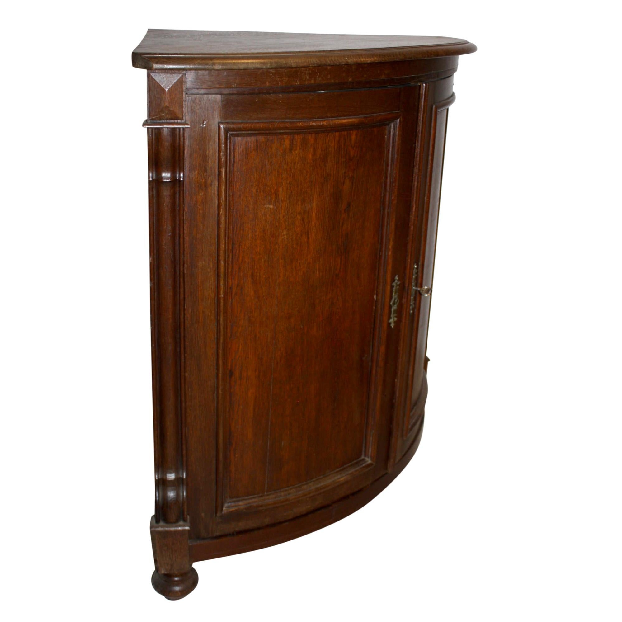 Featuring a bow front with double recessed panel doors and two fixed shelves, this corner cabinet has a wealth of storage. Brass escutcheons with a working Hauenschild key complement the rich patina of the oak. The cabinet rests on turned bun feet.