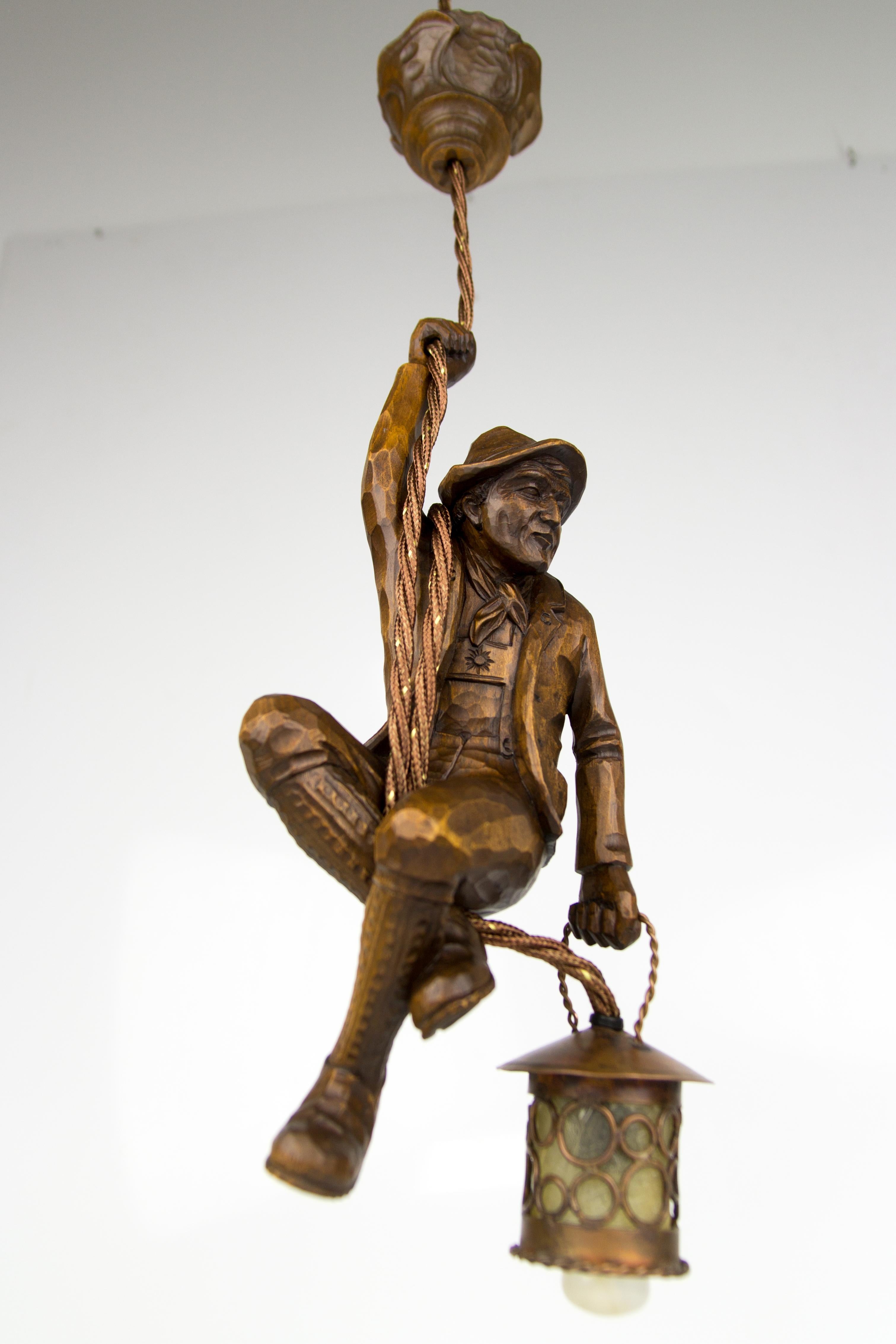 Cette magnifique lampe suspendue figurative de la Forêt Noire allemande présente une figure d'alpiniste sculptée à la main. L'alpiniste en bois sculpté se tient à une corde et tient une lanterne en cuivre dans une main.
La hauteur de la lampe