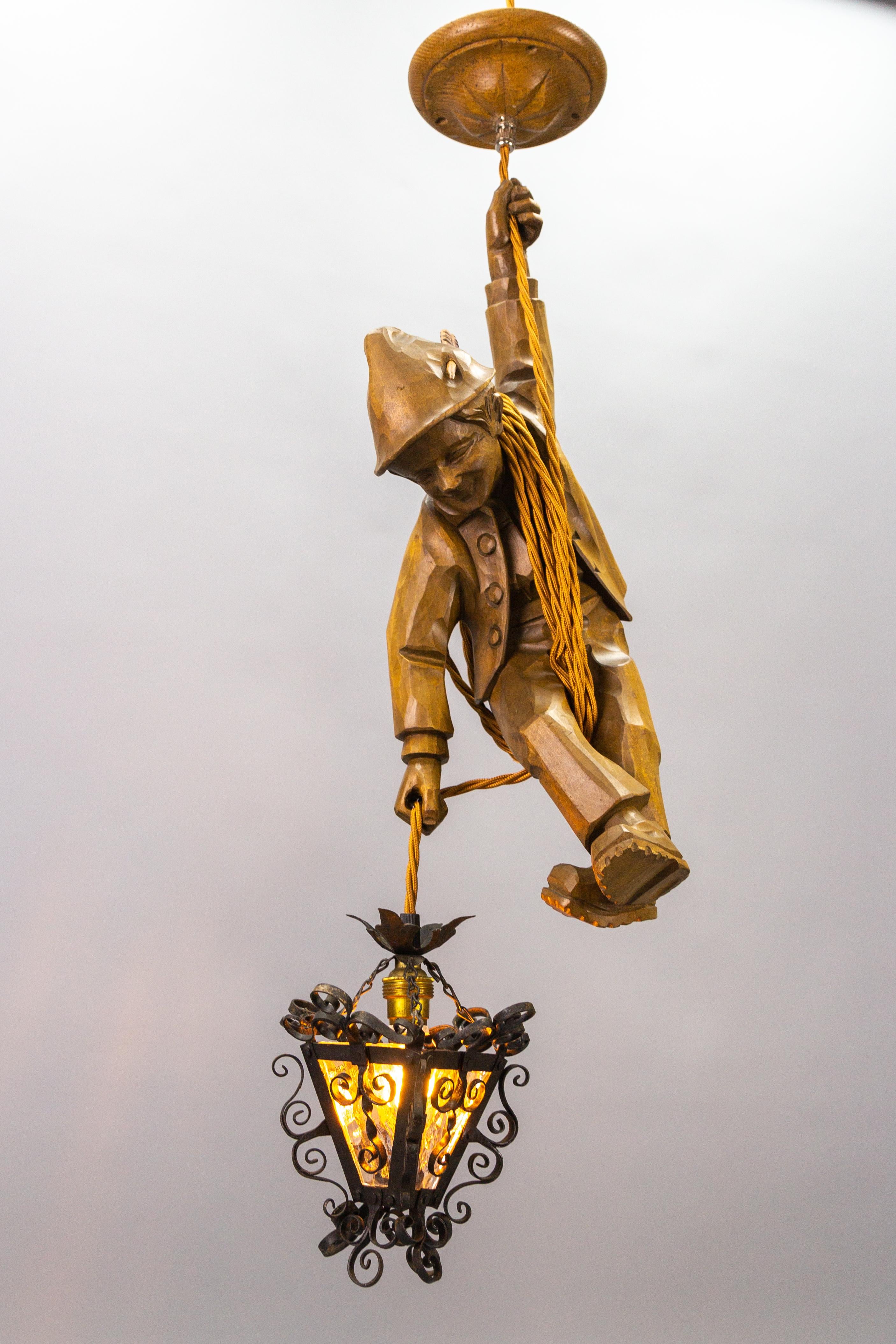 Cette adorable lampe suspendue figurative allemande datant des années 1950 présente une figure sculptée à la main d'un alpiniste souriant. L'alpiniste en bois de tilleul sculpté s'accroche à une corde et tient dans une main une lanterne en métal