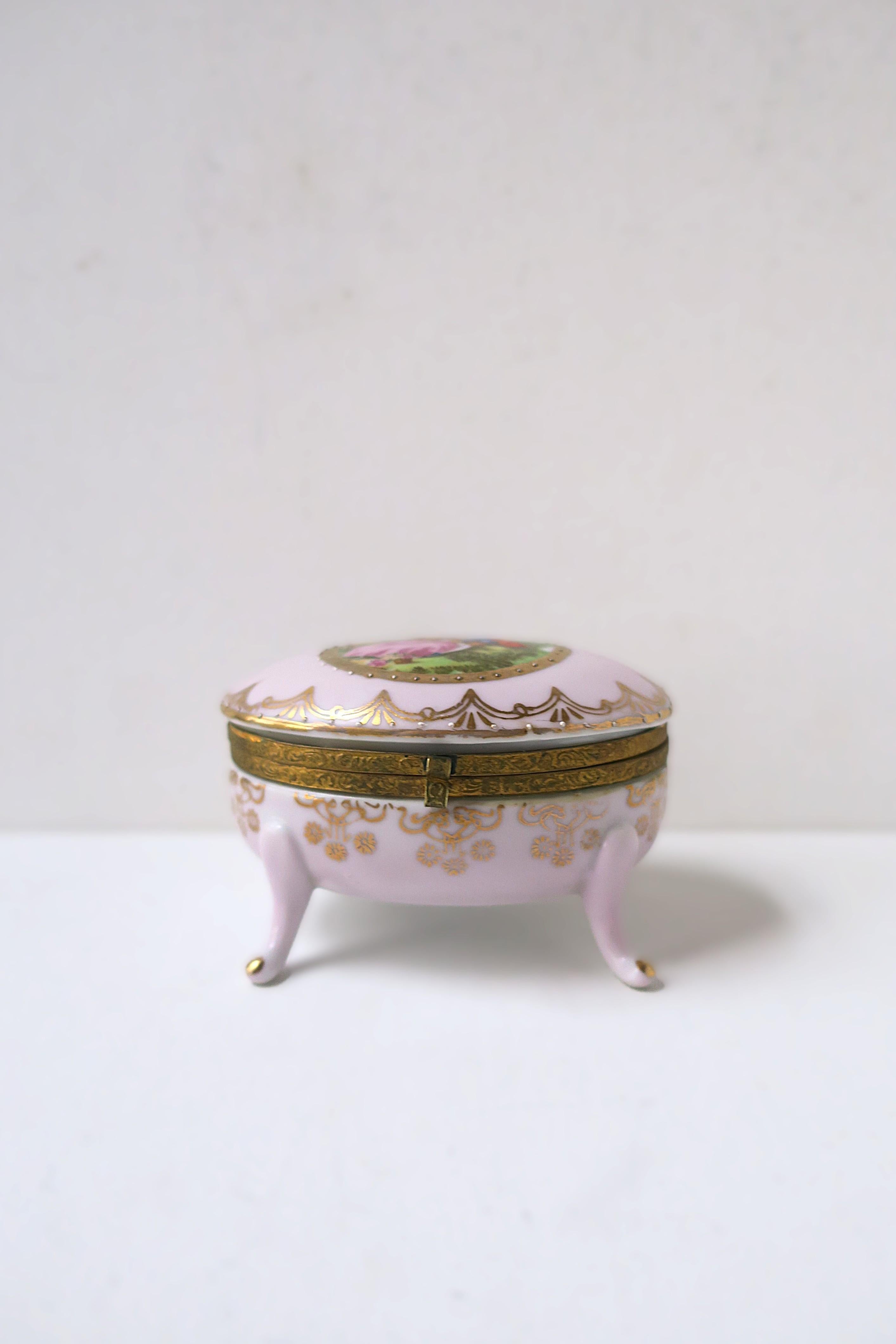 A beautiful German oval porcelain jewelry box, in the Regency design style, circa mid-20th century, Germany. Boxes' lid features outdoor nature scene with couple in a park sounded by trees and flowers with hand-painted gold detail around. Box is