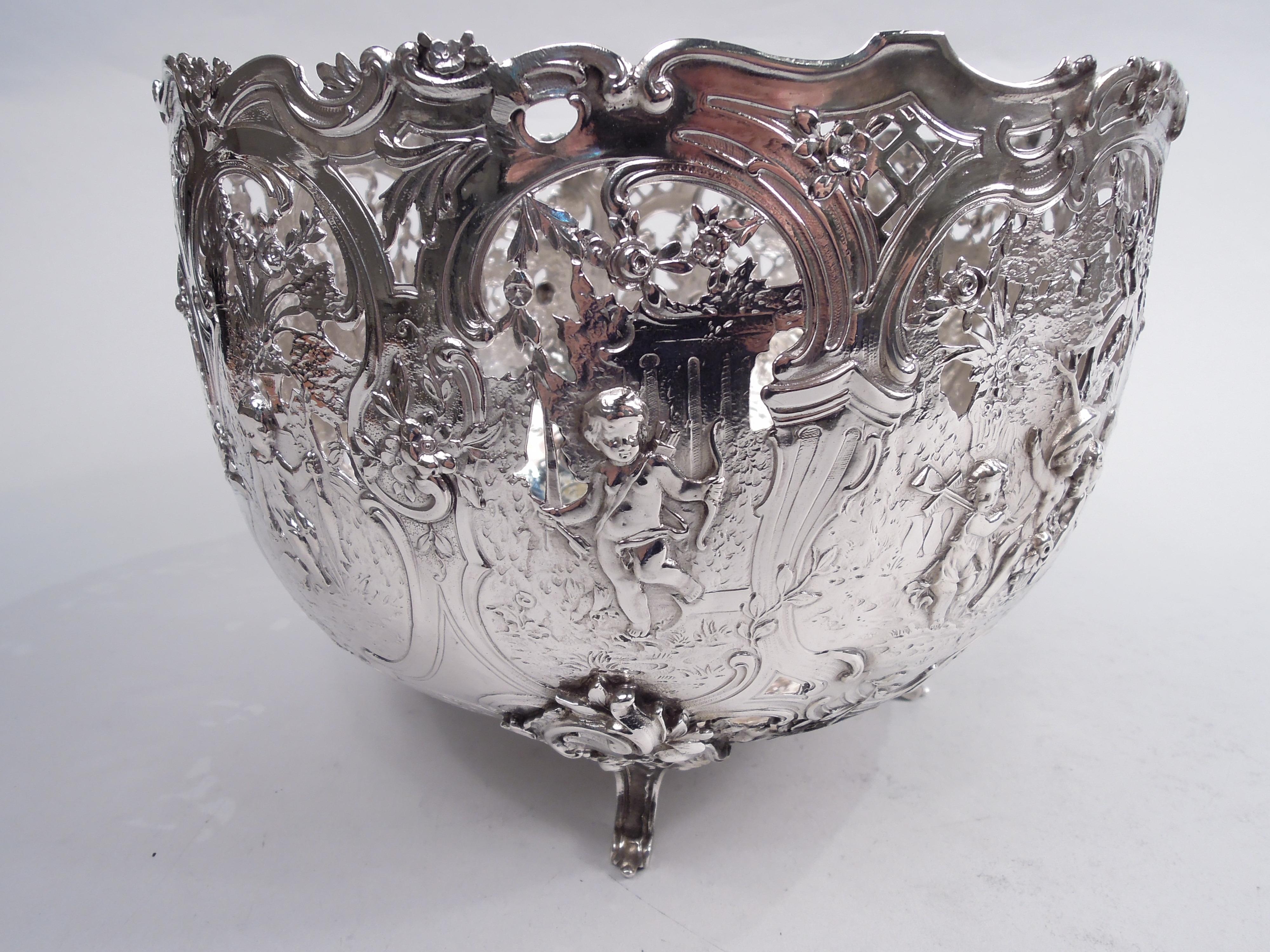German Rococo 800 silver centerpiece bowl, ca 1910. Round and curved with 4 leafing scroll-mounted scroll supports. Chased and open sides with pastoral scenes in scrolled frames depicting cherubs playing at gardening, fishing, and hunting in a