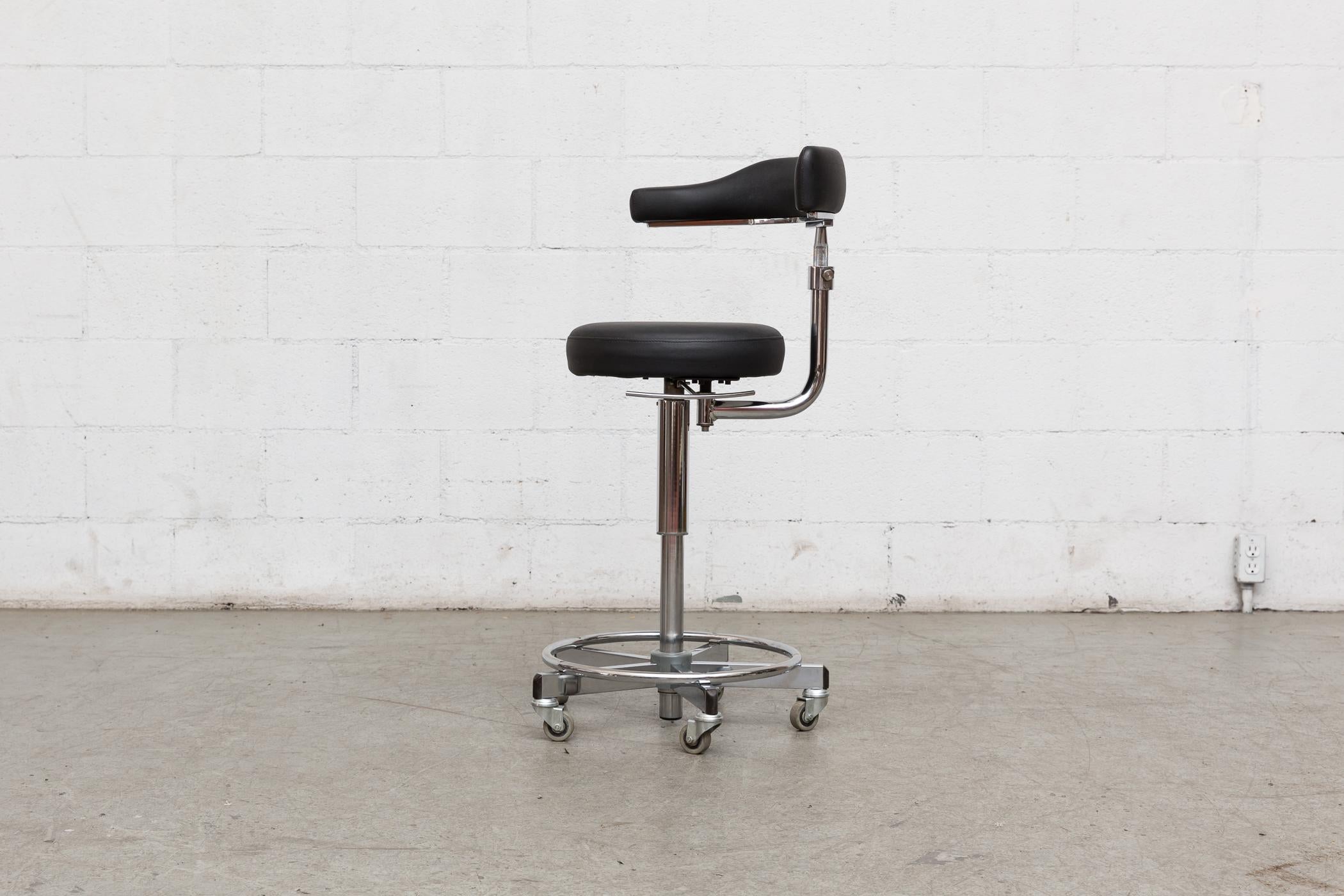 Rolling German dentist chair with asymetric seat back, black leather upholstery and chrome frame with adjustable height. Rubber bumper details on footrest.