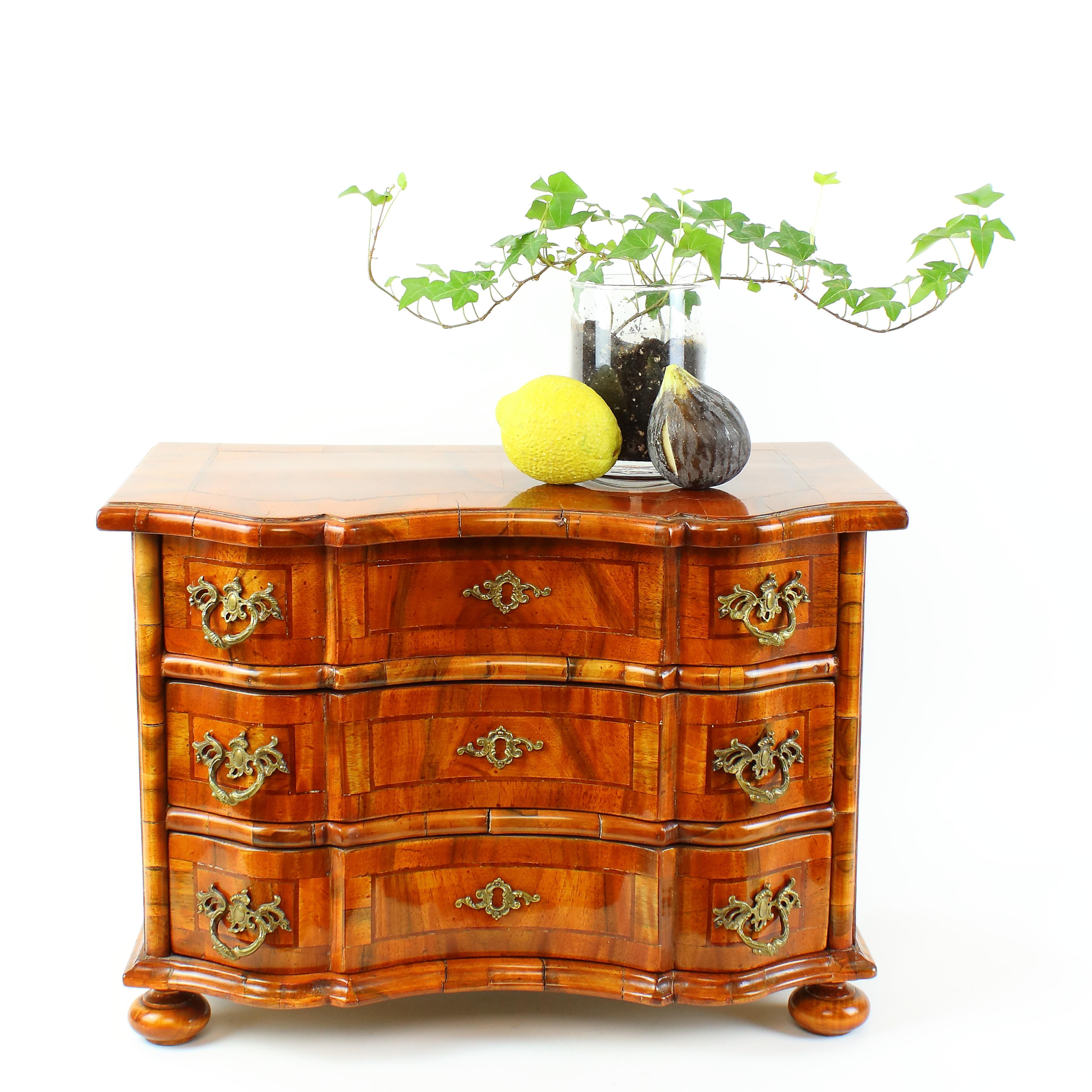 Rectangular German Baroque style chest of drawers with curvy front standing on four bun feet and holding three frieze drawers with bronze handles. Top, front and both sides with fine walnut marquetry. 
The chest of drawers bears all features of a