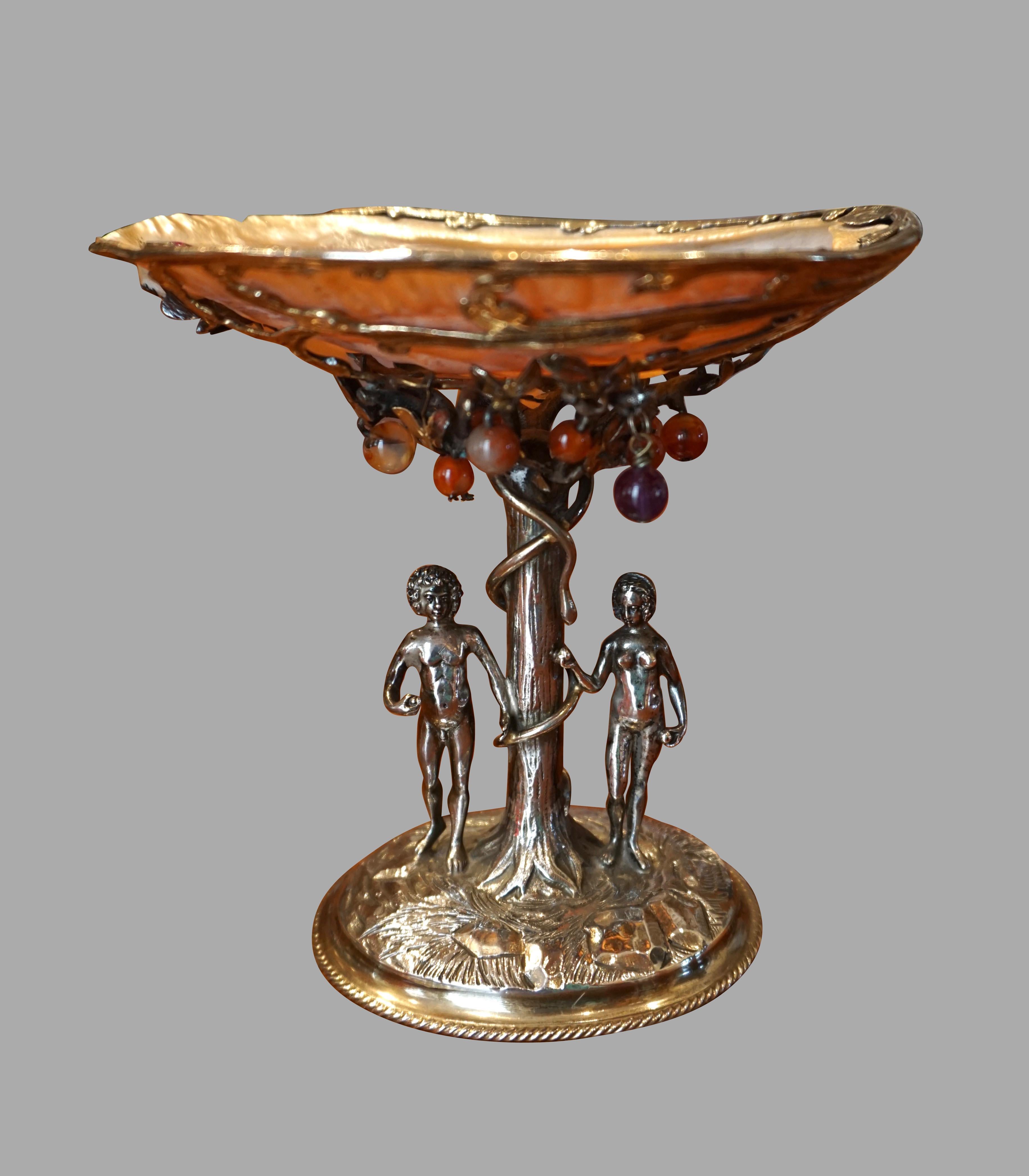 A finely executed silver tazza depicting Adam and Eve in the garden of Eden under the apple tree of knowledge wrapped by a snake. Above their heads is an open abalone shell decorated with semi-precious stones representing apples hanging from the