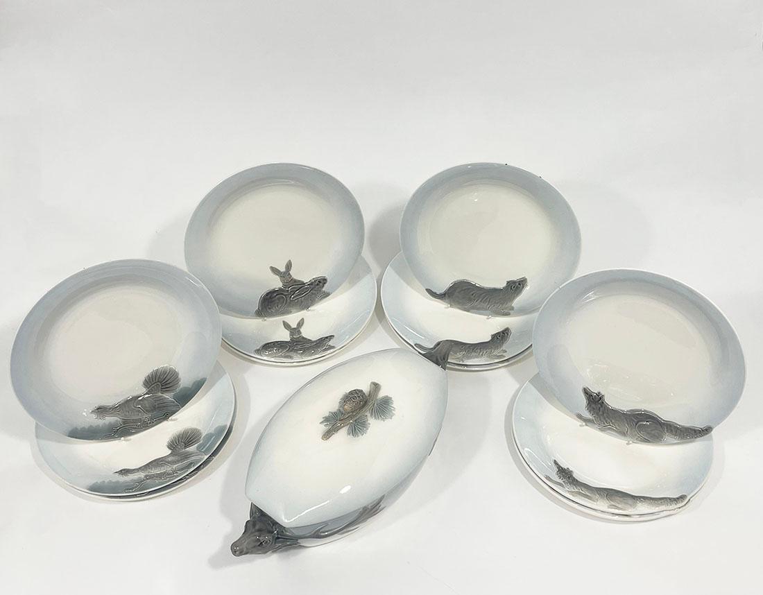 German Wächtersbach 12 Hunting plates and tureen by Carl Bull, 1910

Beige ceramic set with light blue rims and various relief scenes of wild animals in shades of gray. The set consists of a tureen with lid featuring elk head handles and a branch