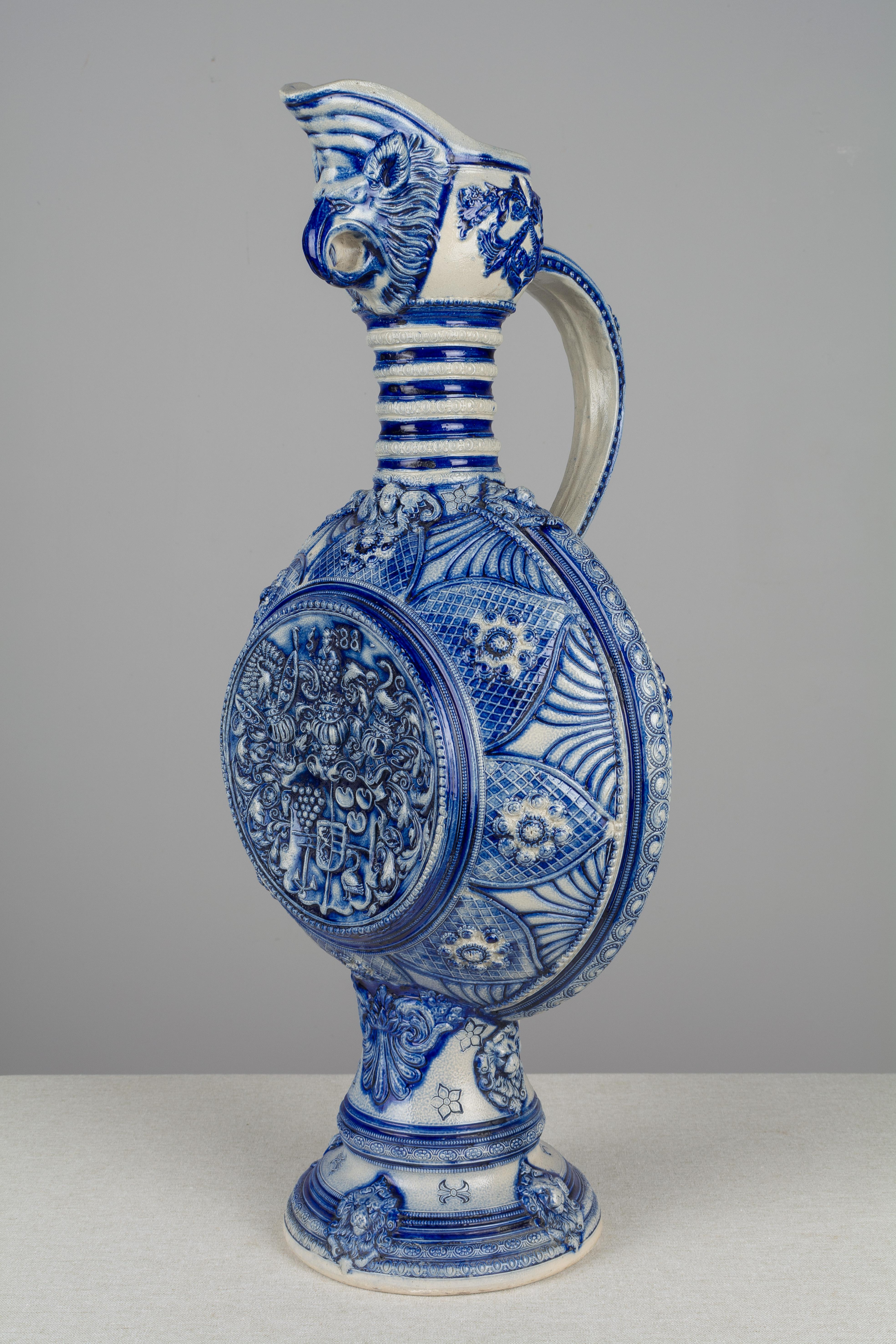 A large German Westerwald pottery ewer with an animal head spout, tall ribbed neck and circular body on a flaring base. Elaborately decorated in deep cobalt blue with heraldic imagery and symbols including a coat of arms, crowns, a peacock and lion
