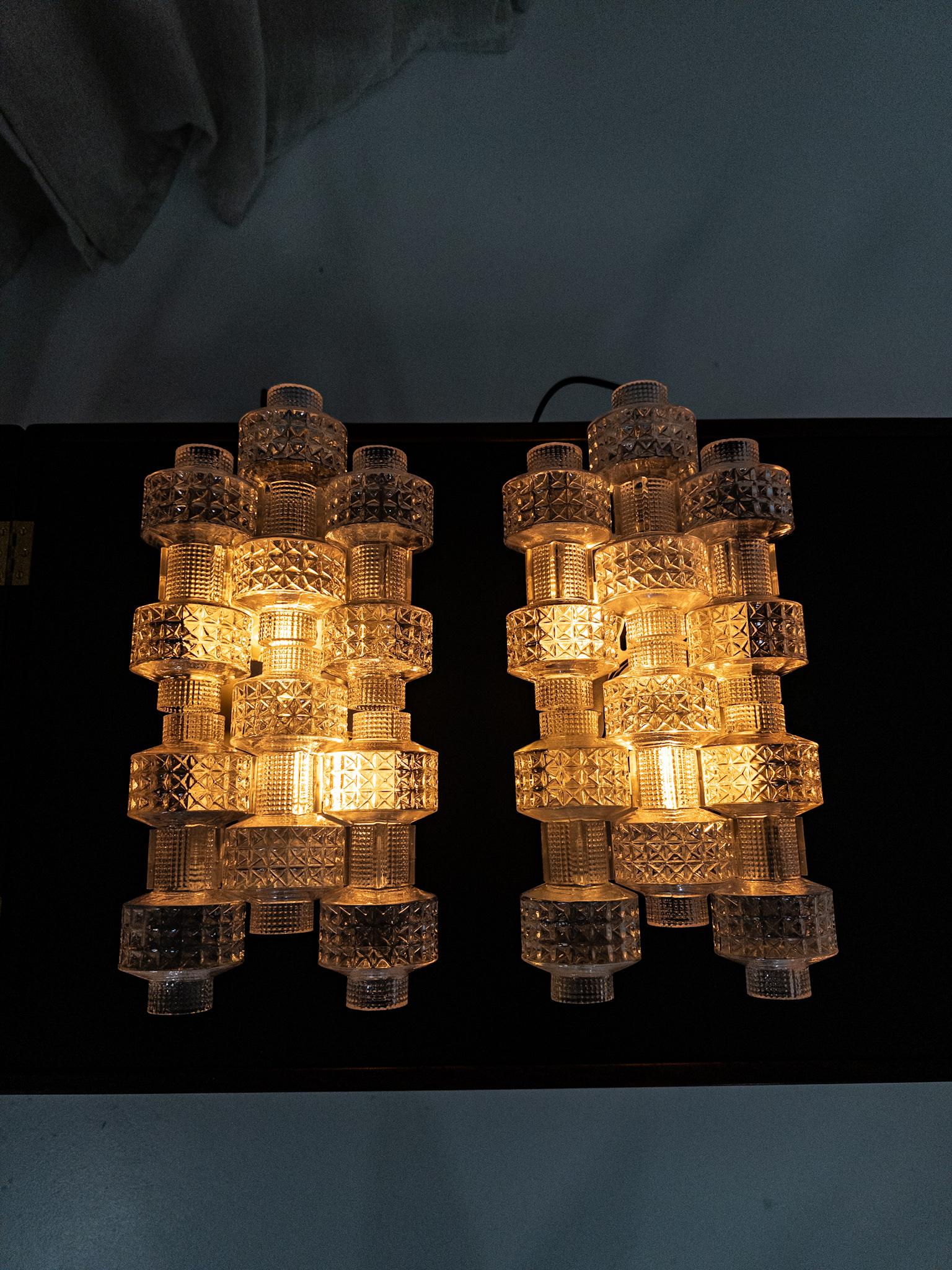 Un par escultórico de apliques de pared Festival compuesto por módulos apilados de cristal prensado texturizado con distintos patrones geométricos. Los gruesos elementos de cristal crean una luz brillante y difusa y confieren a las luminarias su