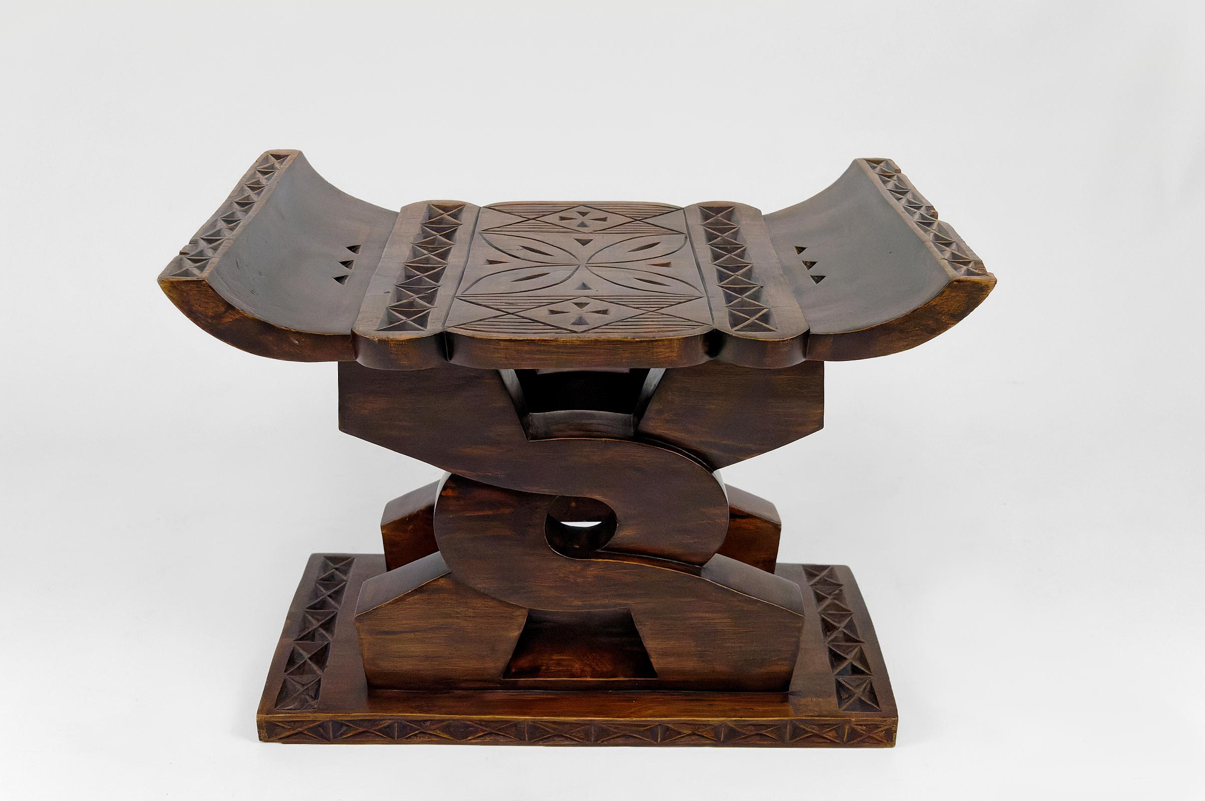 En bois de cèdre.

Tabouret africain en bois sculpté, avec un siège incurvé reposant sur deux supports en bois sculpté entrelacés et une fine base. La tradition ghanéenne explique que le symbole central sculpté dans ce tabouret en cèdre est un nœud