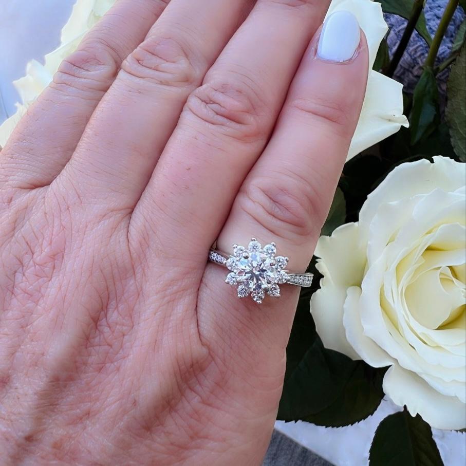 A floral halo twist on a classic design. This stunning 14 karat white gold diamond ring features a 1.00 carat natural round brilliant cut diamond as its centerpiece, surrounded by a halo of fine white diamonds and equally brilliant melee diamonds