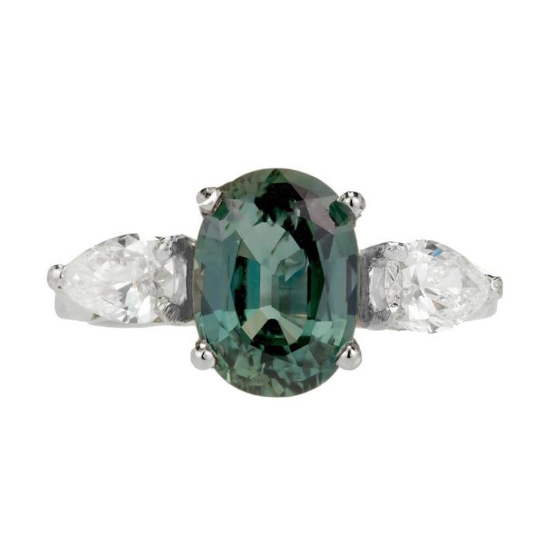 Green Sapphire Diamond Platinum engagement ring. The center stone is a GIA rich blue and green oval sapphire mounted in a platinum setting that is adorned with on pear shaped white diamond on each side. The ring dates back to the 1960's. This
