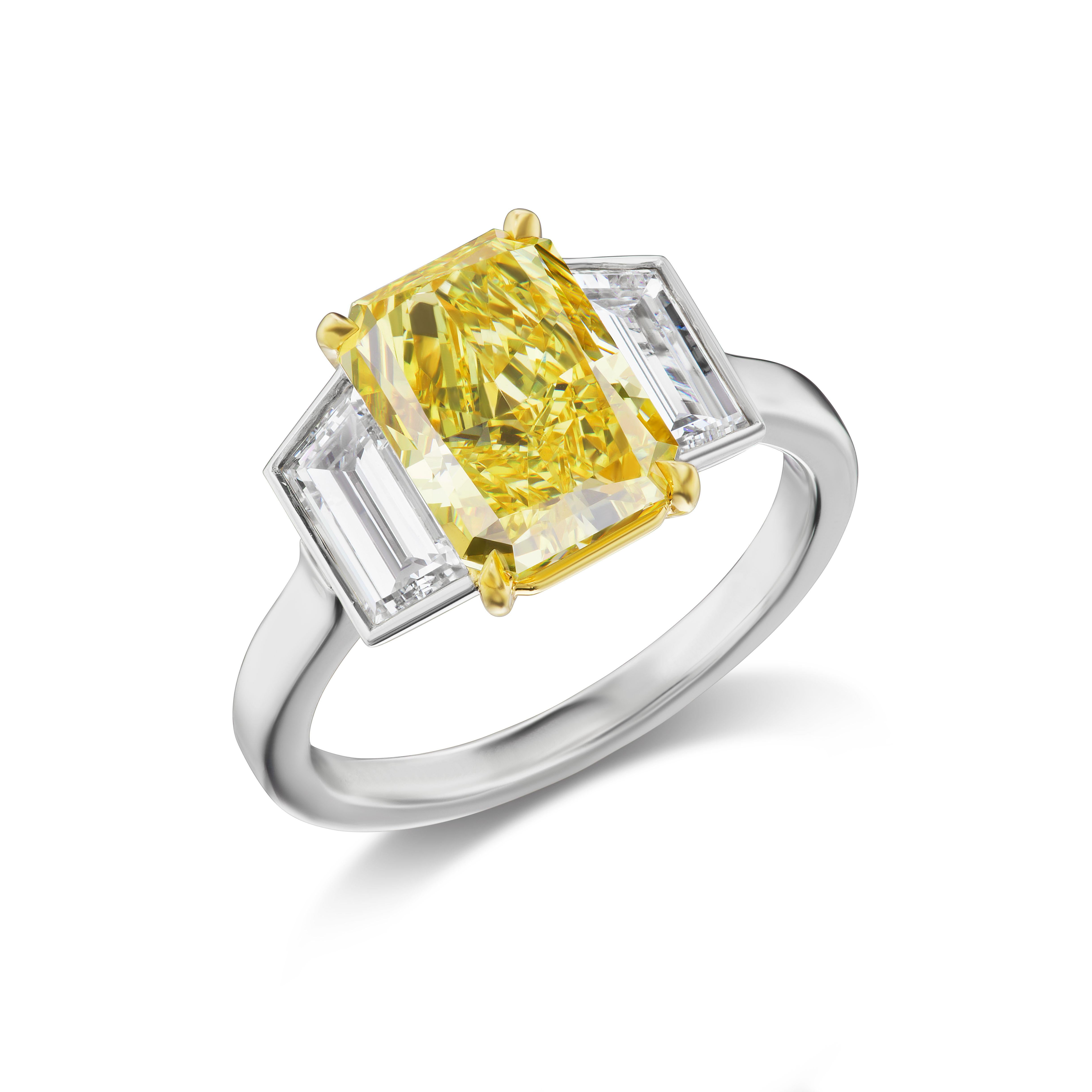 The 3.24 radiant-cut vivid yellow center stone is flanked by 2 trapezoidal diamonds weighing total 1.20 carats in a simple 18K gold and platinum 3 stone ring. The center stone exhibits an exceptional yellow hue, high saturation, and a lovely