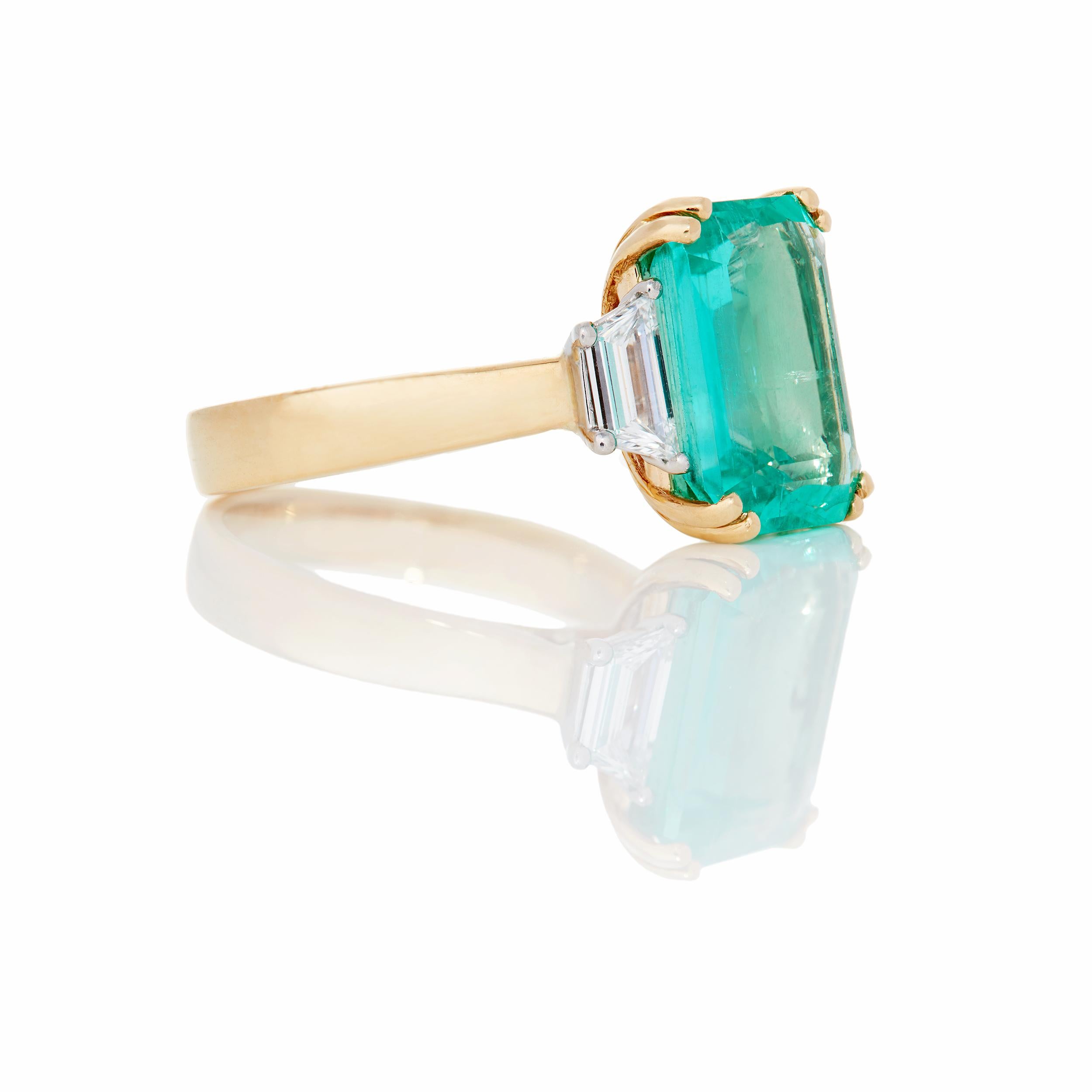 A stunning and ever classic Colombian Emerald ring flanked by two matched Trapezoid Diamonds.  Notice the subtle detail in the profile through use of sweeping lines for an elegant one-of-a-kind ring.  The ring is constructed in 18 Karat Yellow Gold