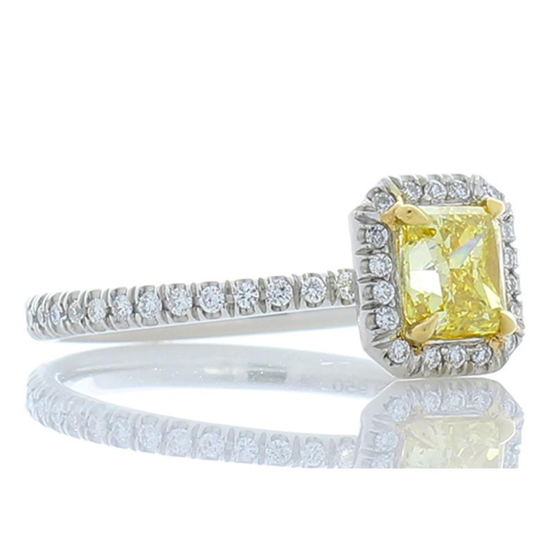 The style of this incredible ring is simple yet sophisticated. It features a dynamic GIA certified 0.73 carat natural fancy intense yellow radiant cut diamond center. Its allure is enhanced by a twinkling halo of bright white diamond melee that