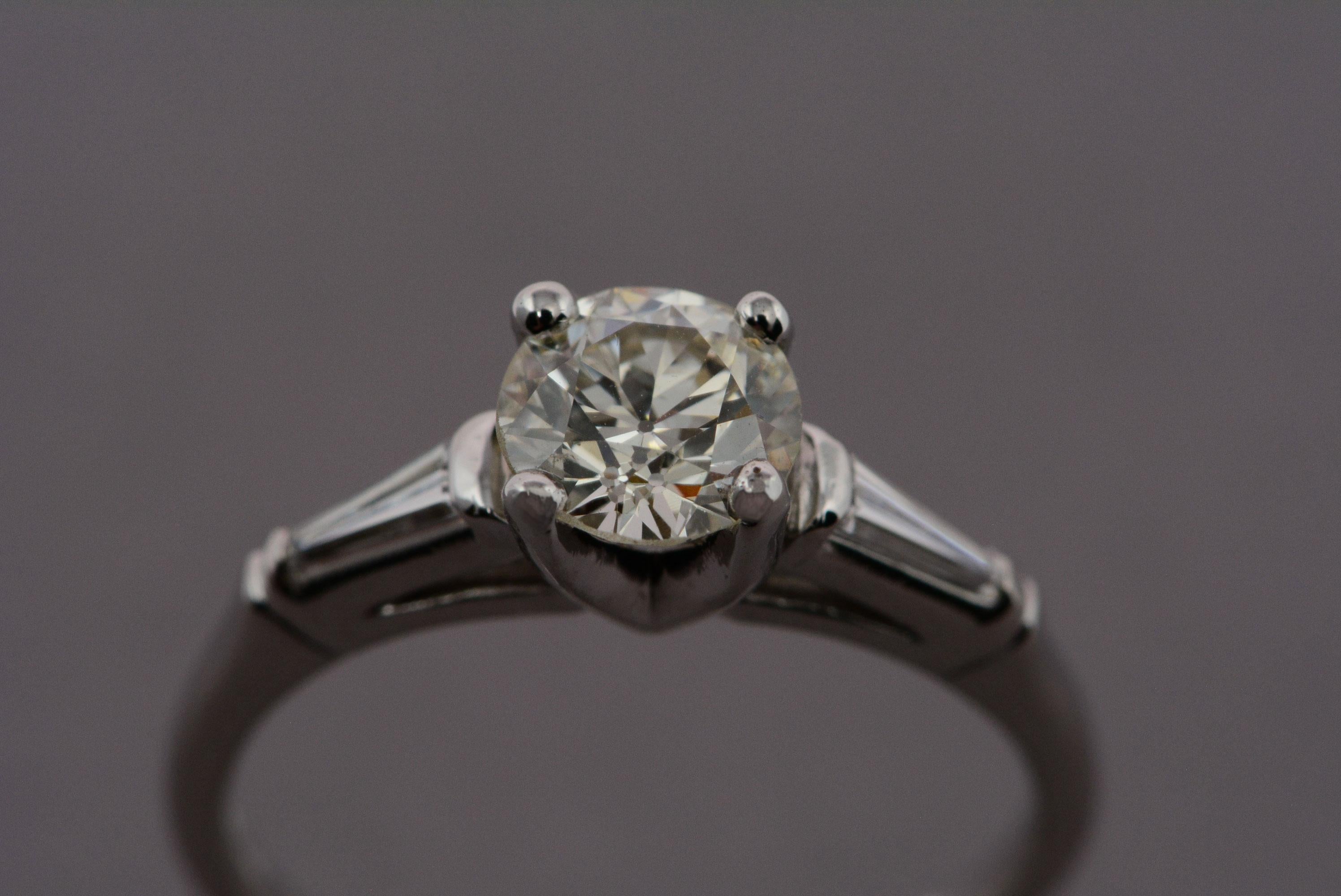 This vintage ring is a classic solitaire style and is made from platinum. 
The ring has two tapered baguette cut diamonds on either shoulder which flow with the tapering of the band. 
The center diamond is a transitional cut, which weighs 1.12