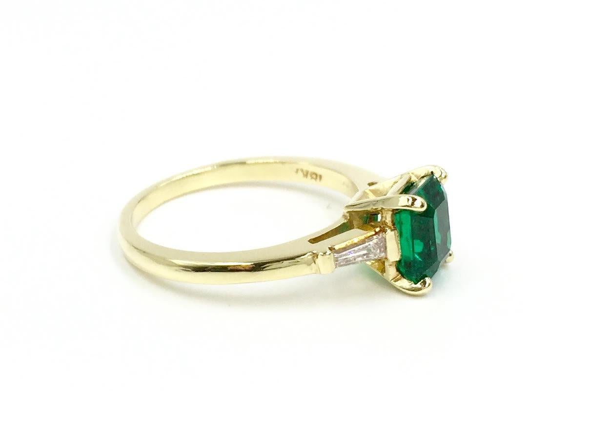 A natural, highly saturated 1.41 carat emerald cut green emerald flanked by bright white slightly tapered baguette diamonds weighing 0.12 carats total weight set in 18 karat yellow gold. High quality green emerald has minimal natural inclusions with