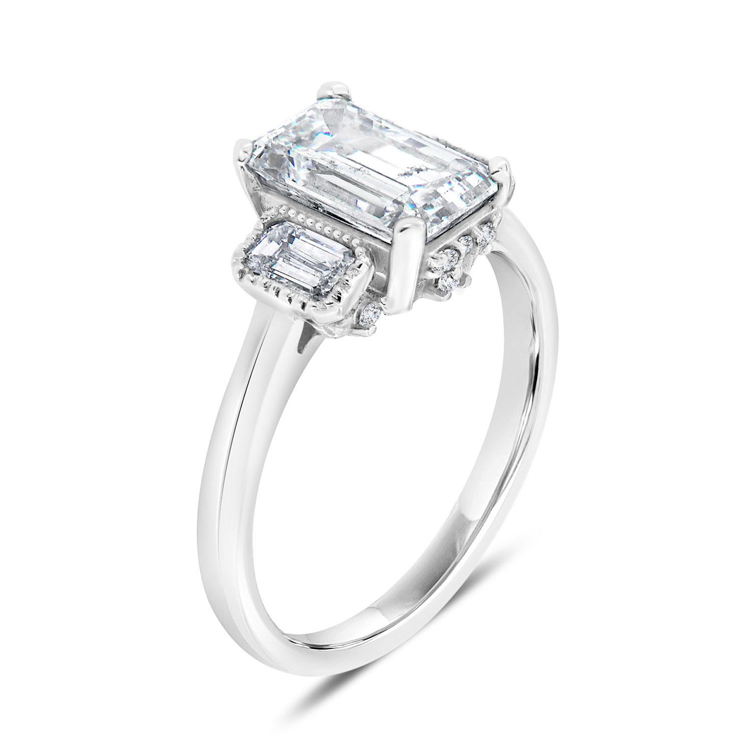 This 14k white gold three-stone ring features a 1.63 carat Emerald cut diamond flanked by two perfectly matched 0.39-carat emerald cut diamonds bezel set with ha delicate Milgrain design around on a 2 mm wide band. We embedded six (6) brilliant