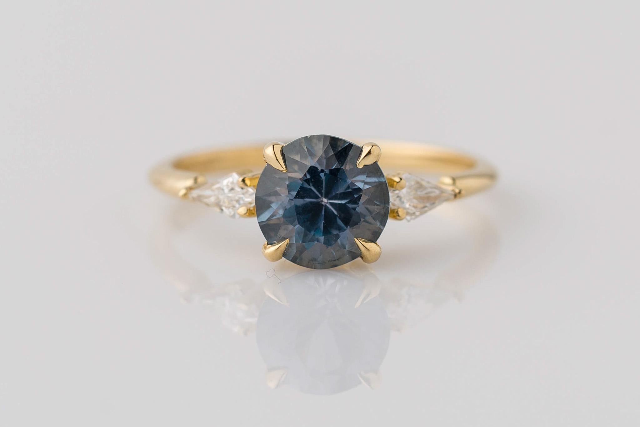 Capture the enchantment of changing hues with our 18K Yellow Gold Montana Color-Changing Sapphire 3-Stone Ring. The centerpiece, an ultra-rare natural untreated Montana sapphire weighing 1.65 carats, exhibits a mesmerizing shift from gray-blue to