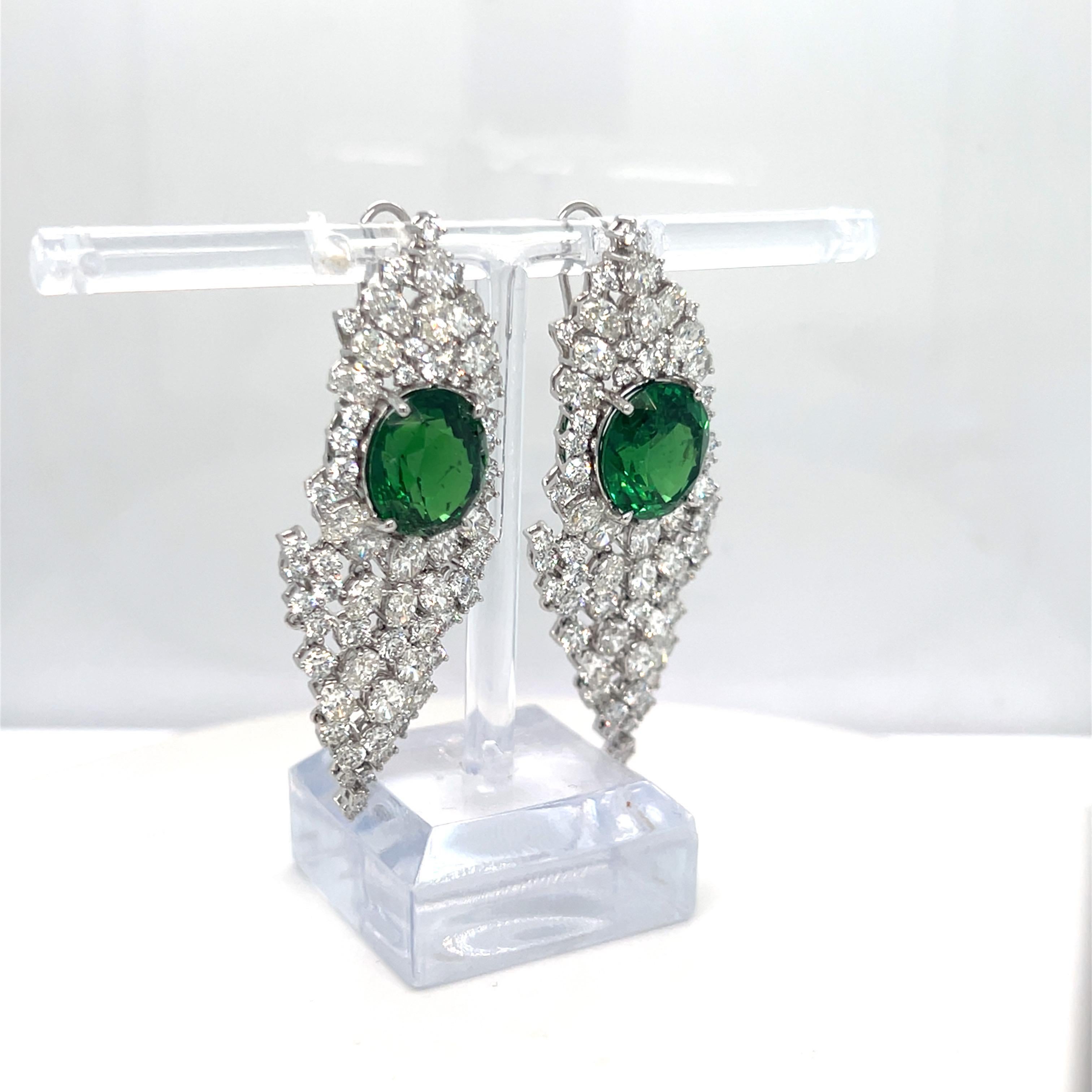 The earrings are set in a timeless 18K white gold setting, designed to showcase the vibrant 17.97-carat tsavorite diamond at the center. The surrounding 7.18-carat oval and 4.21-carat round white diamonds are meticulously arranged in a cluster,