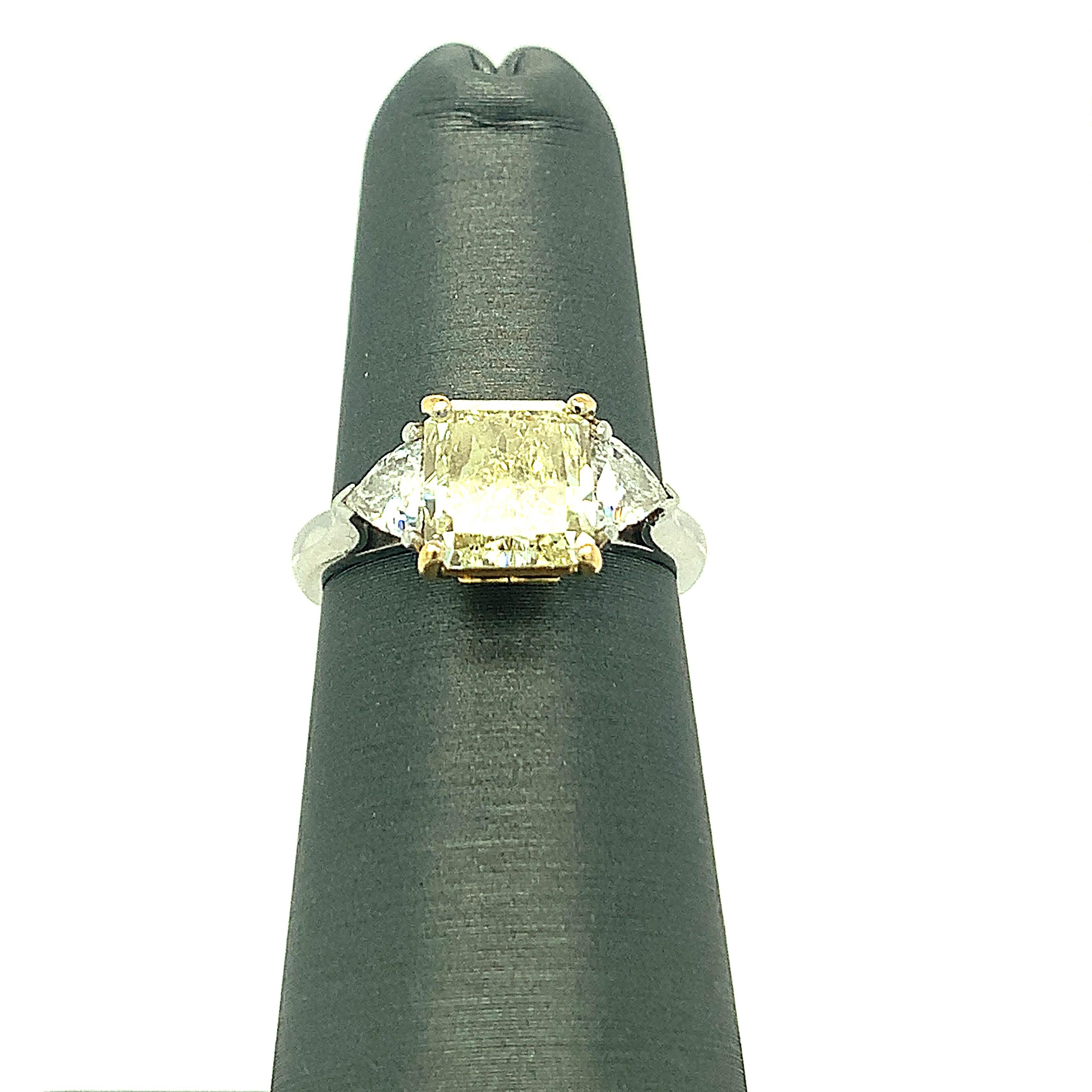 1.83 Carat Cornered Square cut Fancy Light Yellow Diamond is accompanied with a GIA certificate. This brilliant center stone sits on 18K yellow gold mount with platinum band. Two perfectly matched white trillion cut diamonds on sides enhances its