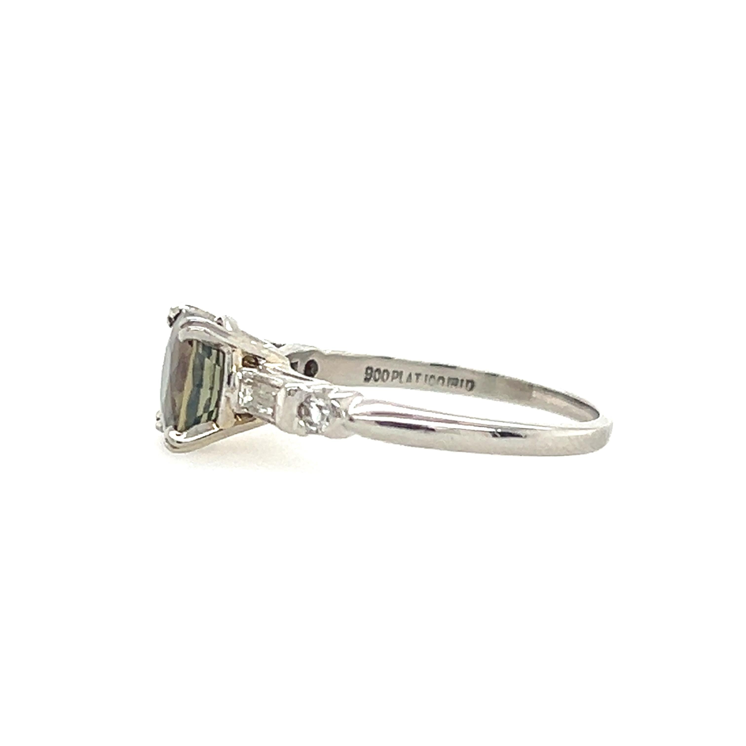 This exquisite 1950s platinum ring showcases a rare 1.87 carat cushion cut natural alexandrite, a gemstone celebrated for its remarkable color-changing properties. Alexandrite famously shifts from greenish hues in daylight to shades of red or purple