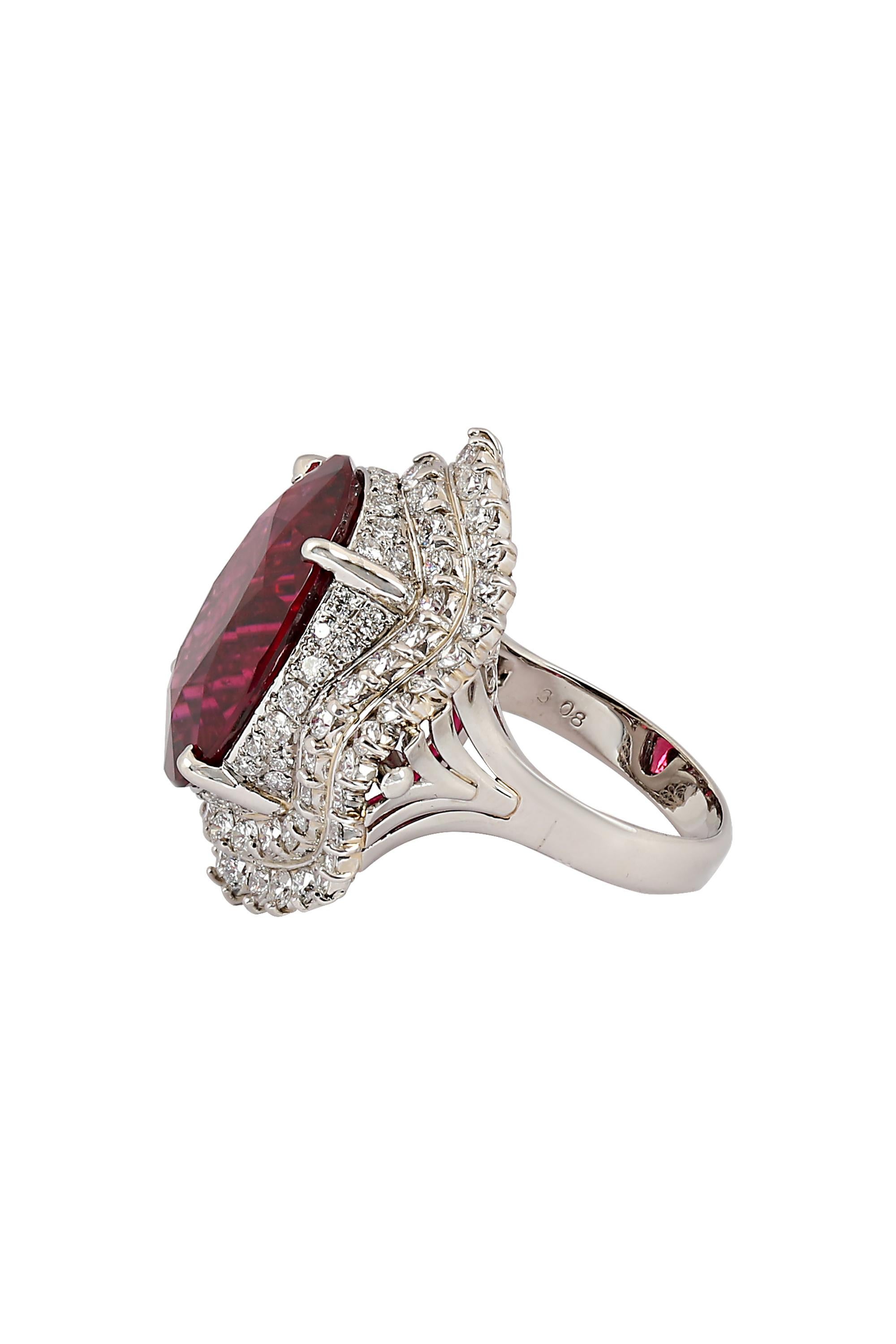 A rich and regal red faceted oval Rubellite weighing 21.85 carats radiates from within the center of an elaborate three-tiered frame of round brilliant diamonds. A scrollwork undercarriage completes the finely crafted 18-karat white gold mounting.