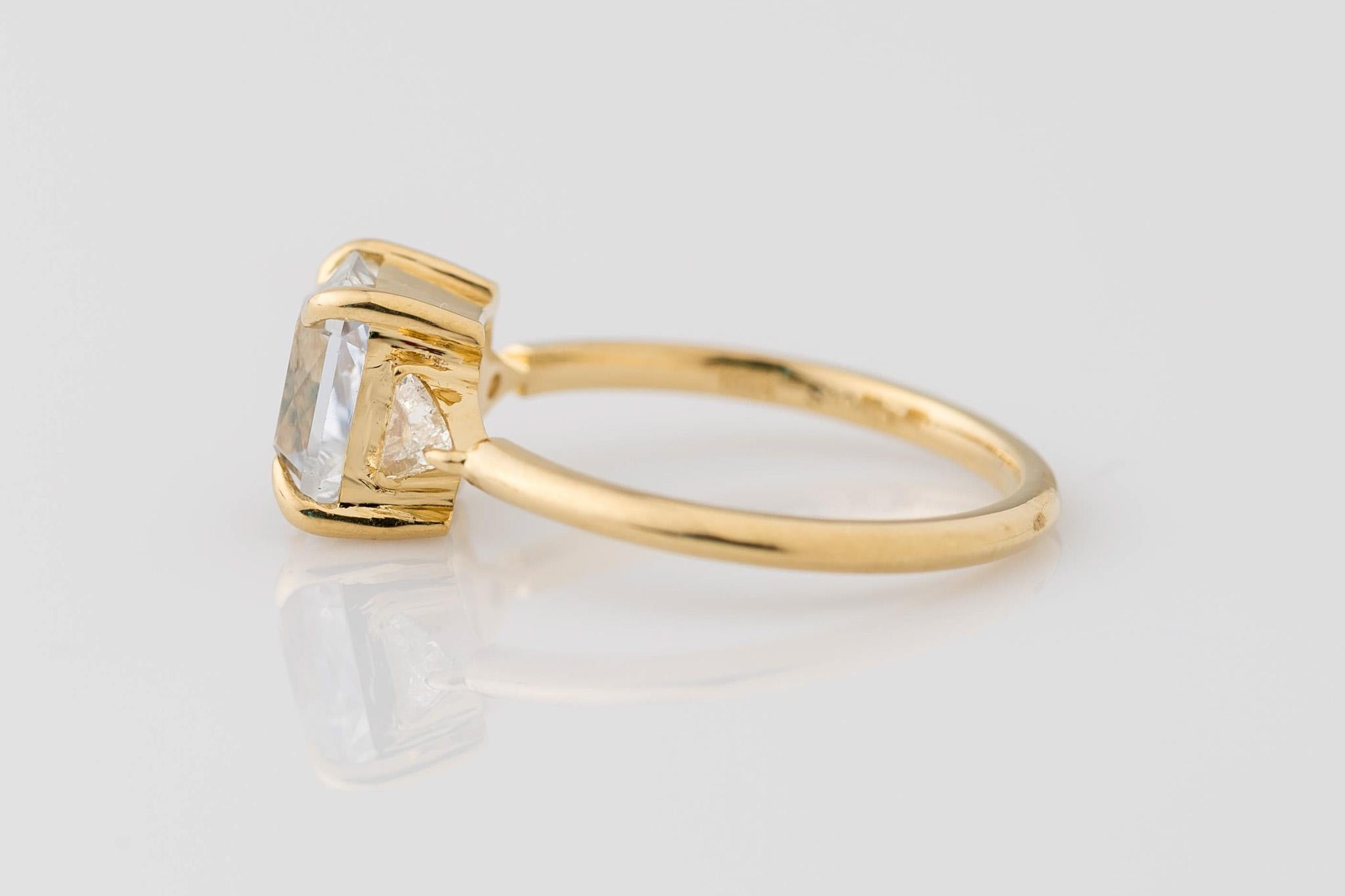 Celebrate your love with our exquisite 18K Yellow Gold GIA Certified Radiant Cut White Sapphire Engagement Ring, accentuated with Trillion Diamonds. The centerpiece boasts a GIA certified 2.22 carat white sapphire, measuring 7.25x6.35x5.16MM, in its