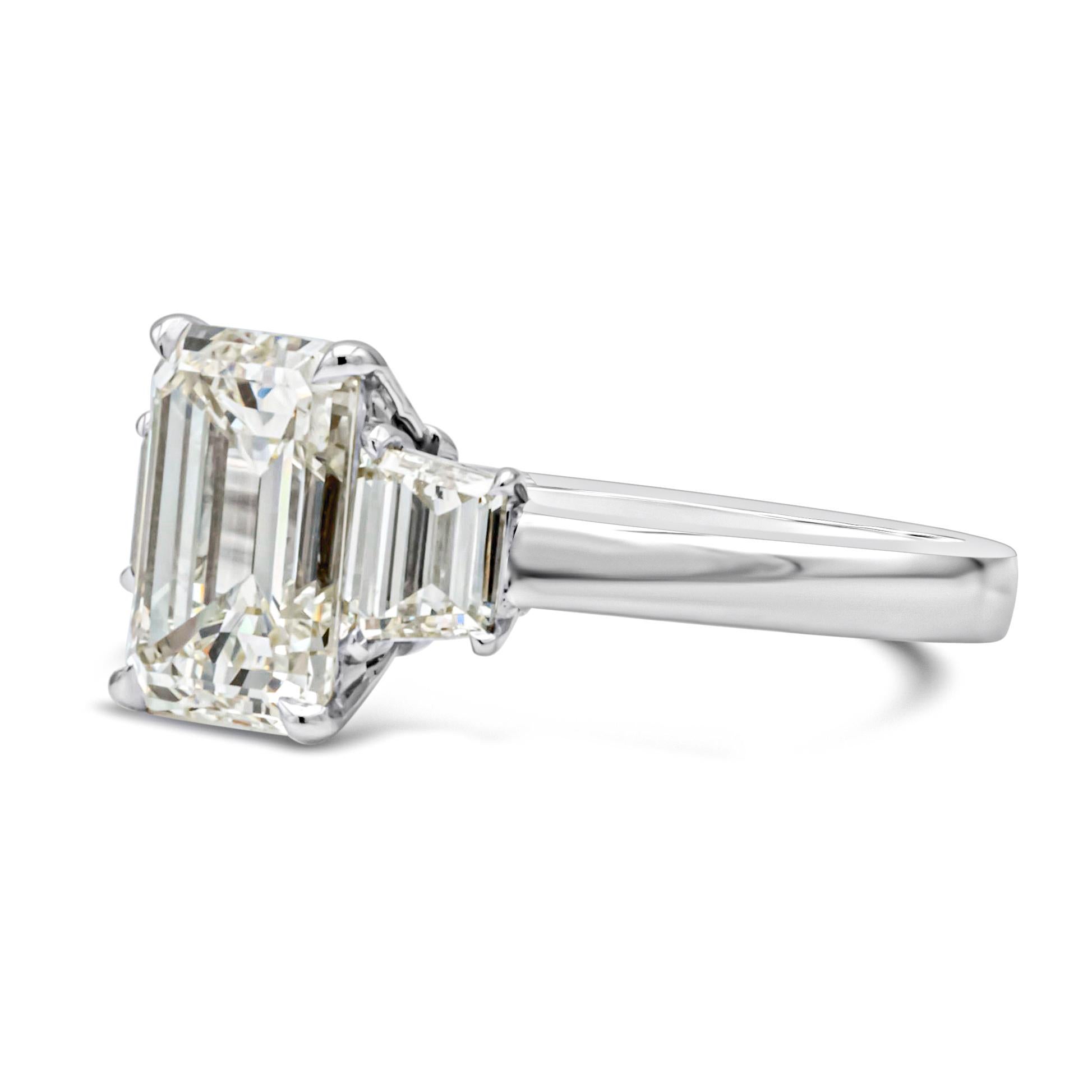 A magnificent three-stone engagement ring, showcasing a 2.96 carats emerald cut diamond certified by GIA as M color and VS1 clarity, set on a four prong setting. Flanked by two perfectly matching trapezoid cut diamonds weighing 0.70 carat total with