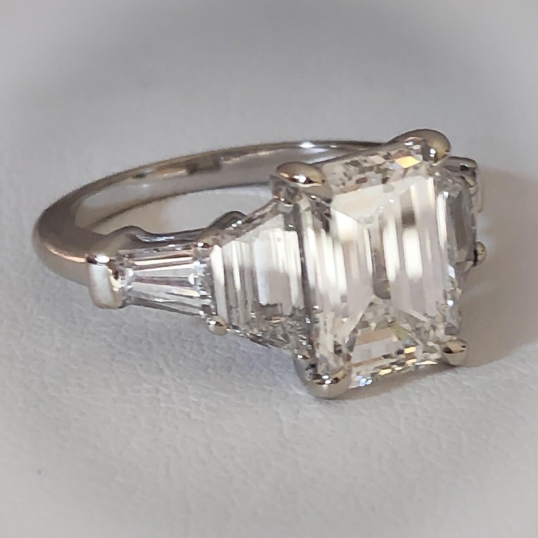 This ring is designed in 14 karat white gold and set with a stunning 3.03 carat GIA certified Emerald Cut diamond. The center diamond is a fancy emerald step-cut shape, H color, VVS1 clarity. It is flanked by two white trapezoid step-cut diamonds