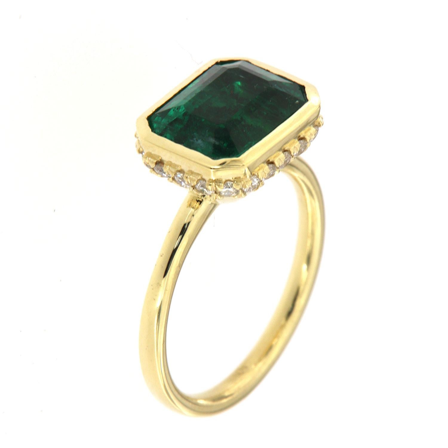 This 18k yellow gold delicate ring features a 3.93 Carat Vibrant green Zambian emerald cut Emerald bezel set East-West style. Twenty-one (21)Brilliant round diamonds are micro-prong set in a hidden halo on the crown to create the sparkle look every