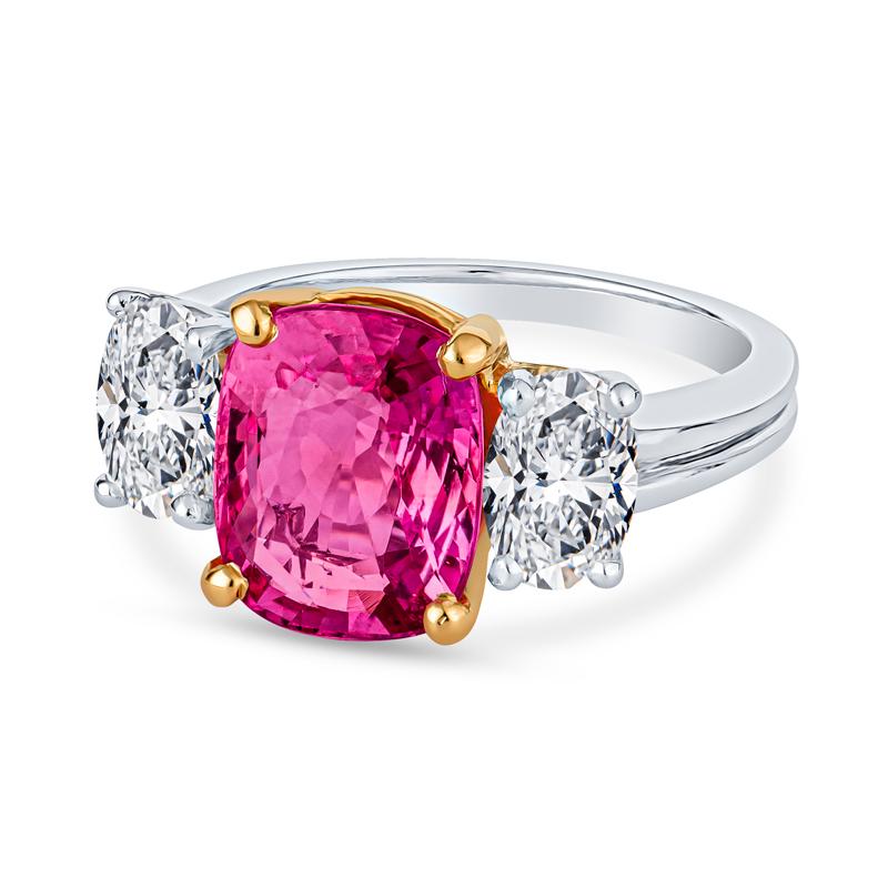 This rare 3.98ct cushion cut natural Padparadscha sapphire from Madagascar, certified by GIA, is the centerpiece of this stunning 3-stone ring. The orangey pink gemstone is set in 18 karat yellow gold and beautifully complemented by two oval shaped
